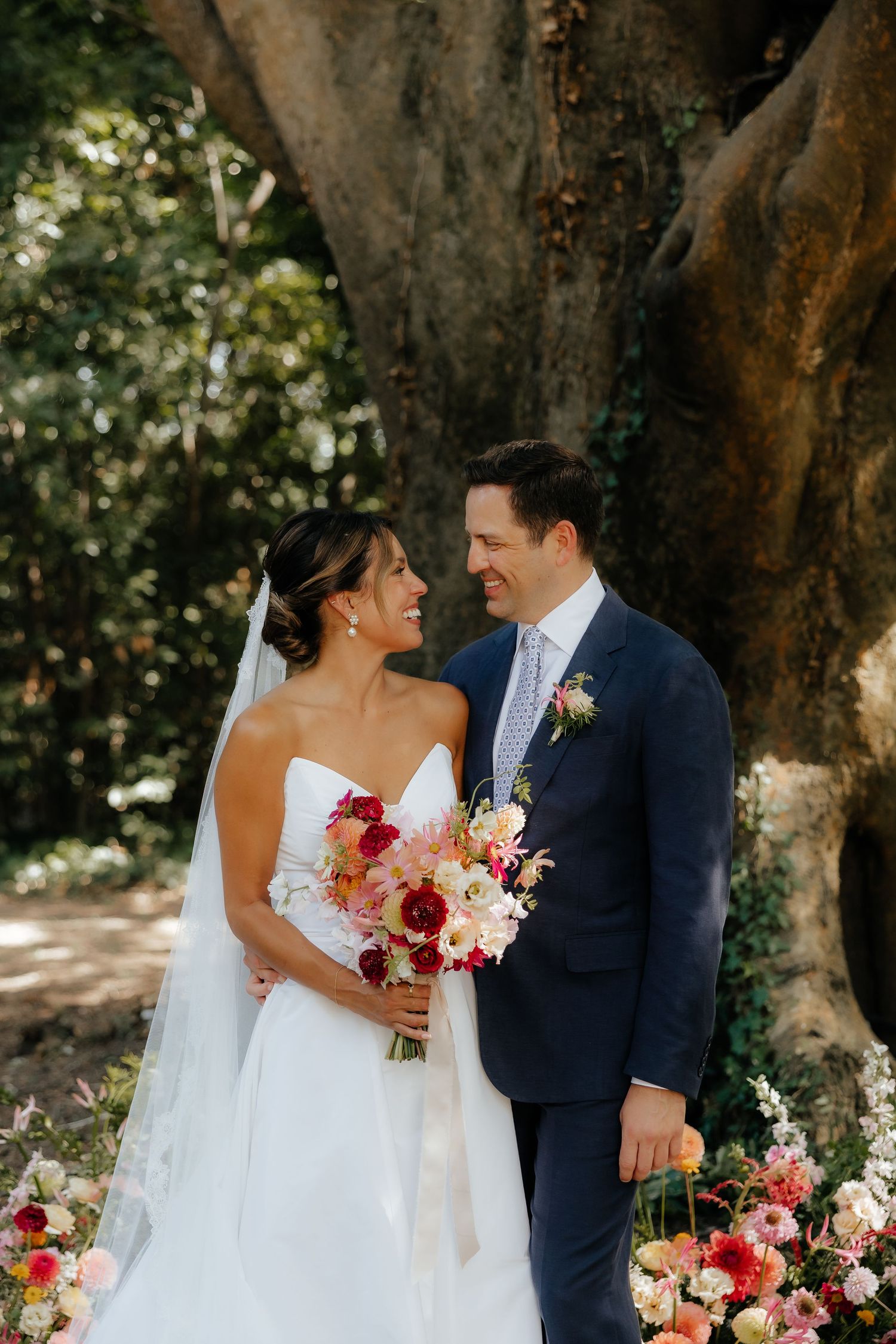 A wedding couple poses under a large tree with pink and red floral bouquet arrangements.