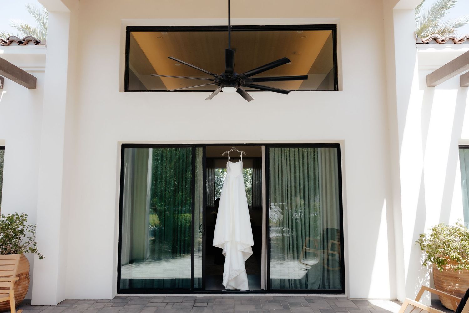 Elegant white wedding gown hanging in doorway with ceiling fan and lush greenery visible outside.