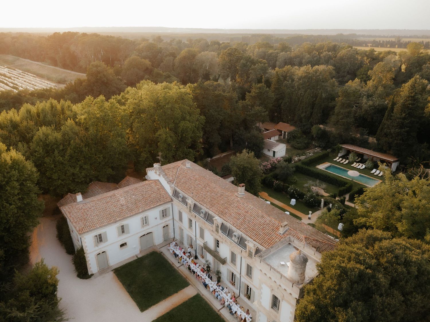 Aerial panoramic view of a large white villa estate surrounded by lush gardens and trees at sunset.