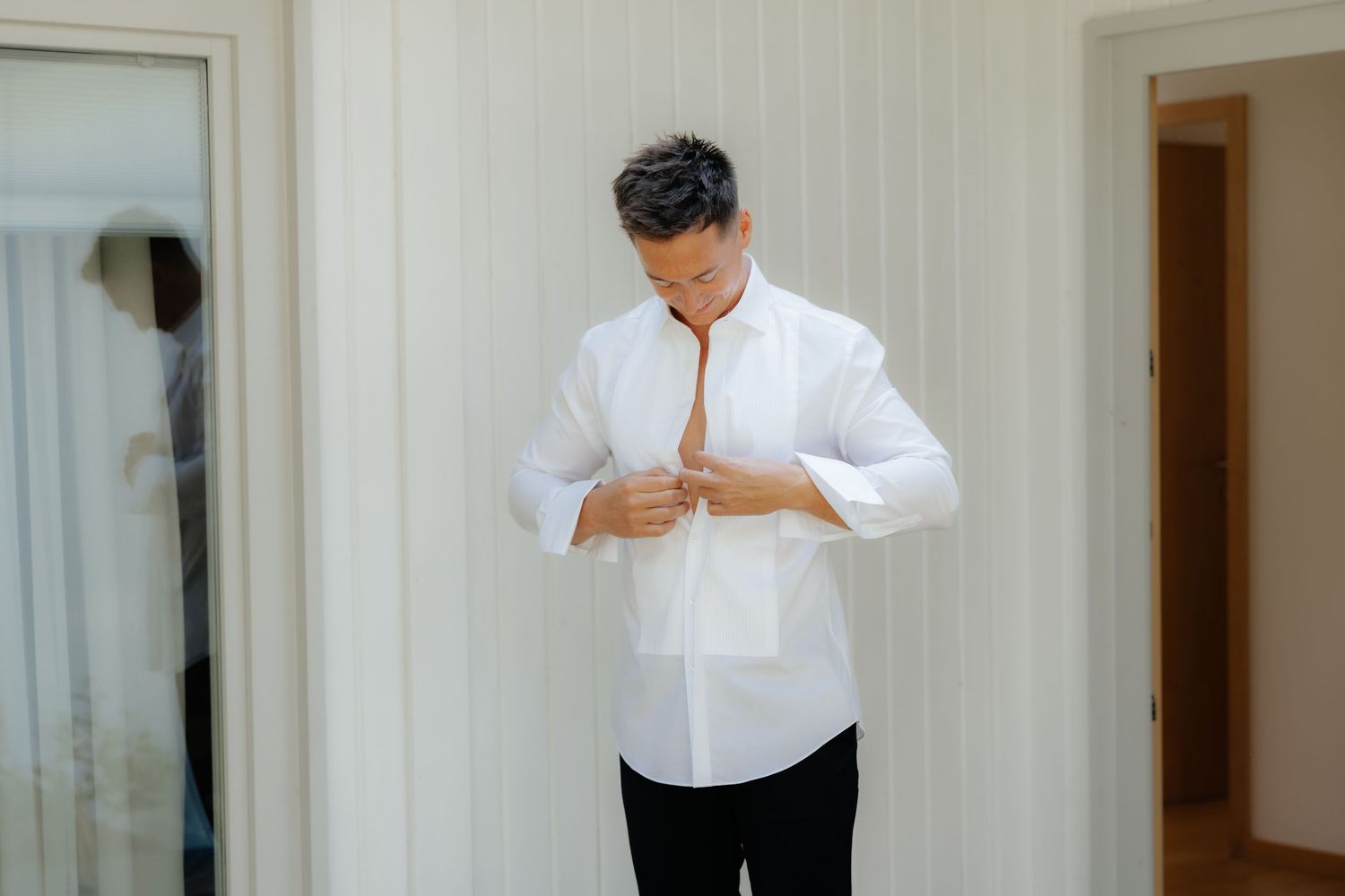 Someone in a white dress shirt and black pants buttons up their shirt while getting dressed in a bright room.