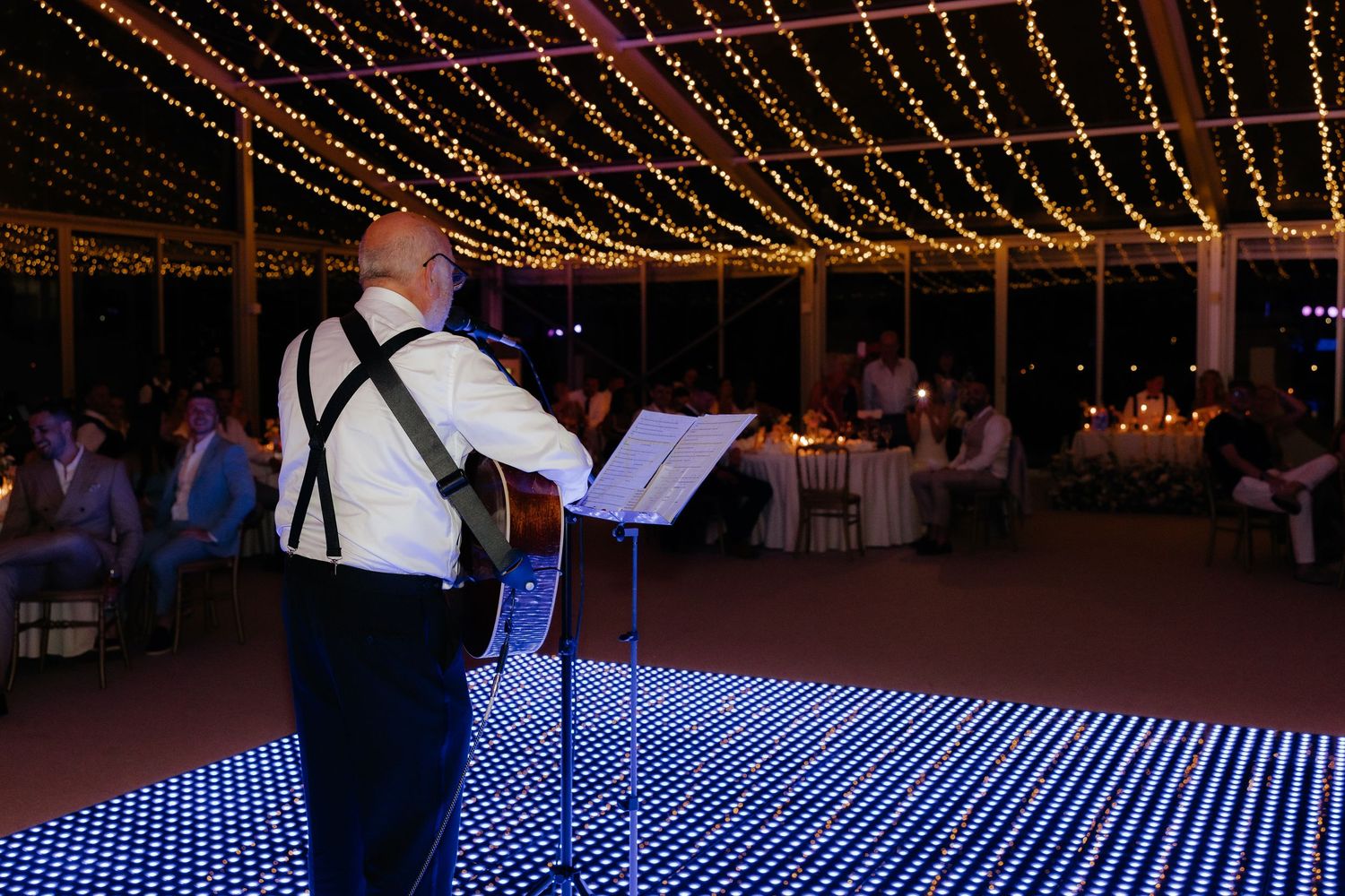 A performer stands on an illuminated LED dance floor under string lights at an evening reception.