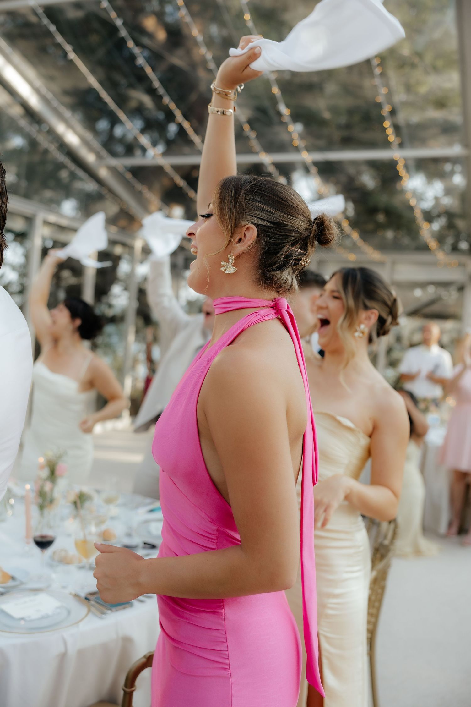 A group of wedding guests in pink and white dresses laughing and celebrating at an outdoor reception.