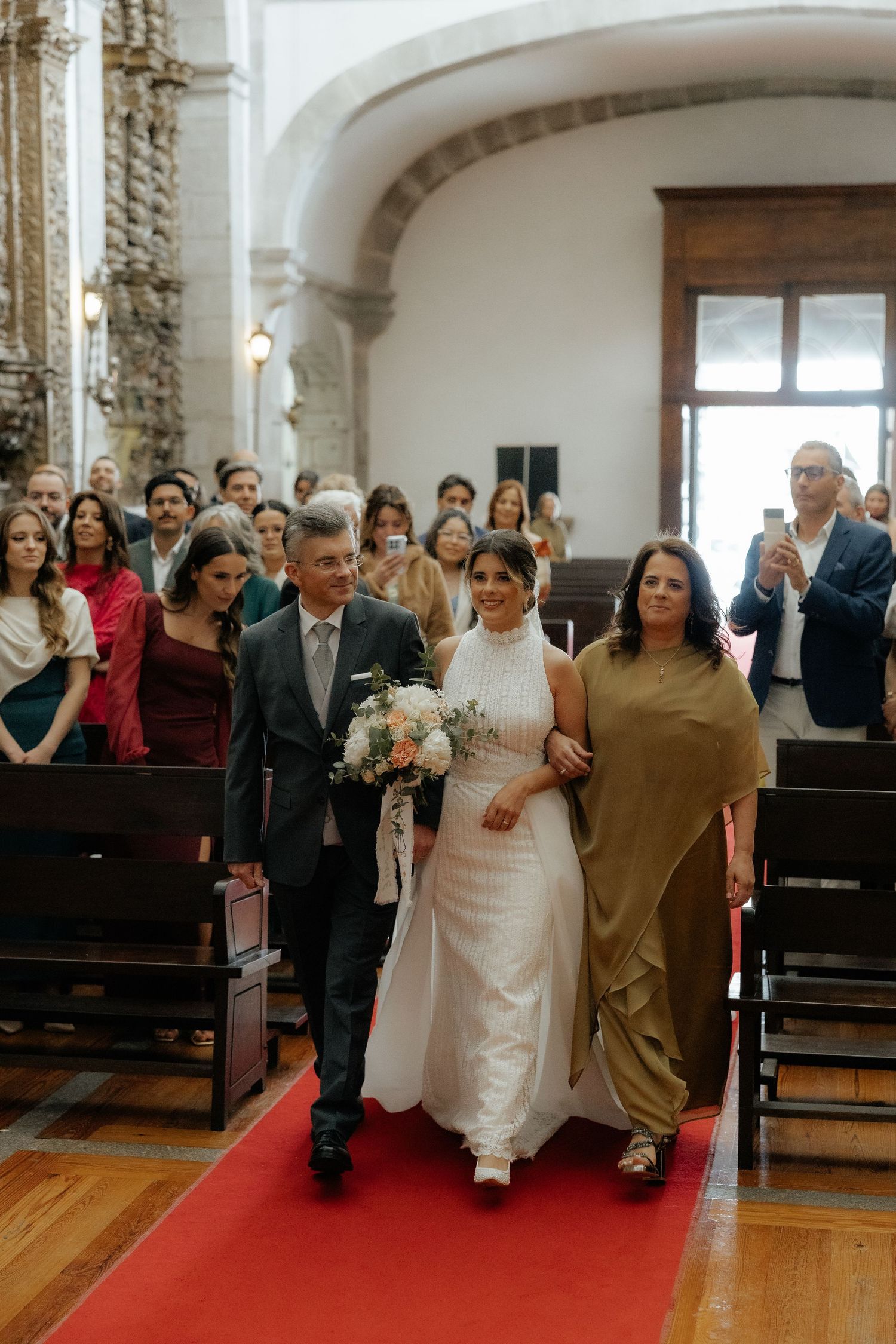 Bride walks down red carpet aisle escorted by parents in beautiful stone church.