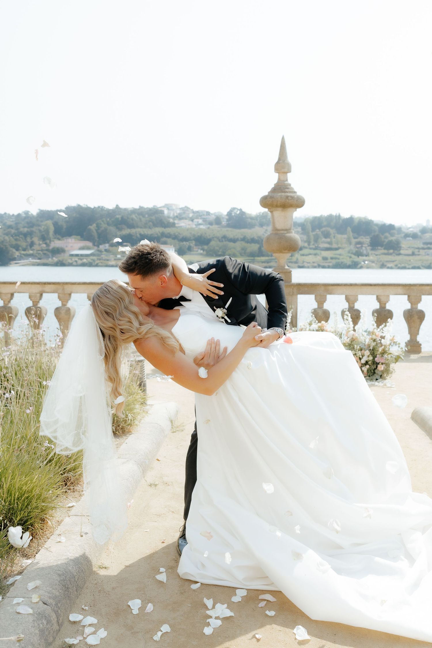 Groom dips bride dramatically on sunlit terrace with stone balustrade and river backdrop at wedding.