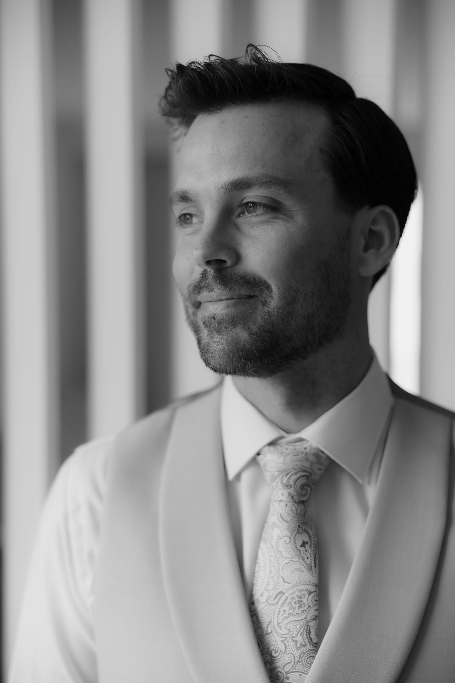 Black and white portrait of handsome groom in white vest and paisley tie gazing thoughtfully.