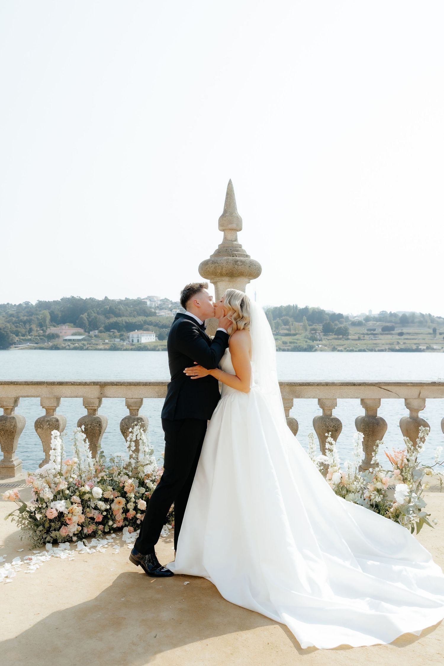 Romantic wedding couple sharing a kiss on a scenic stone balcony overlooking the water with floral arrangements.