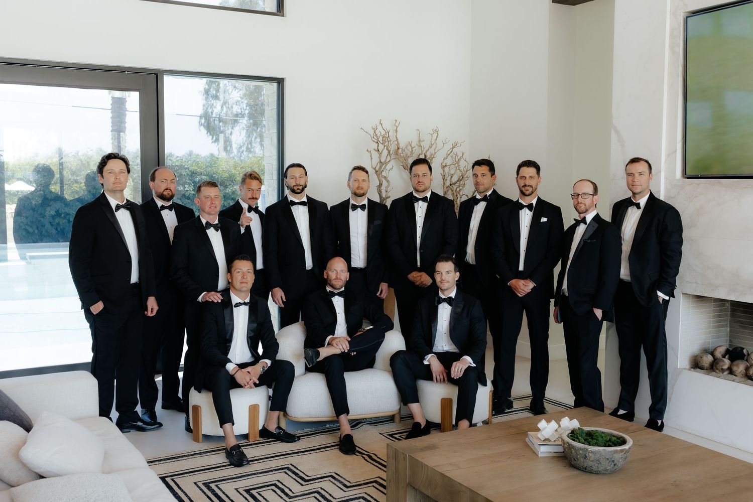 Large group of groomsmen posing in matching black tuxedos in modern white interior.