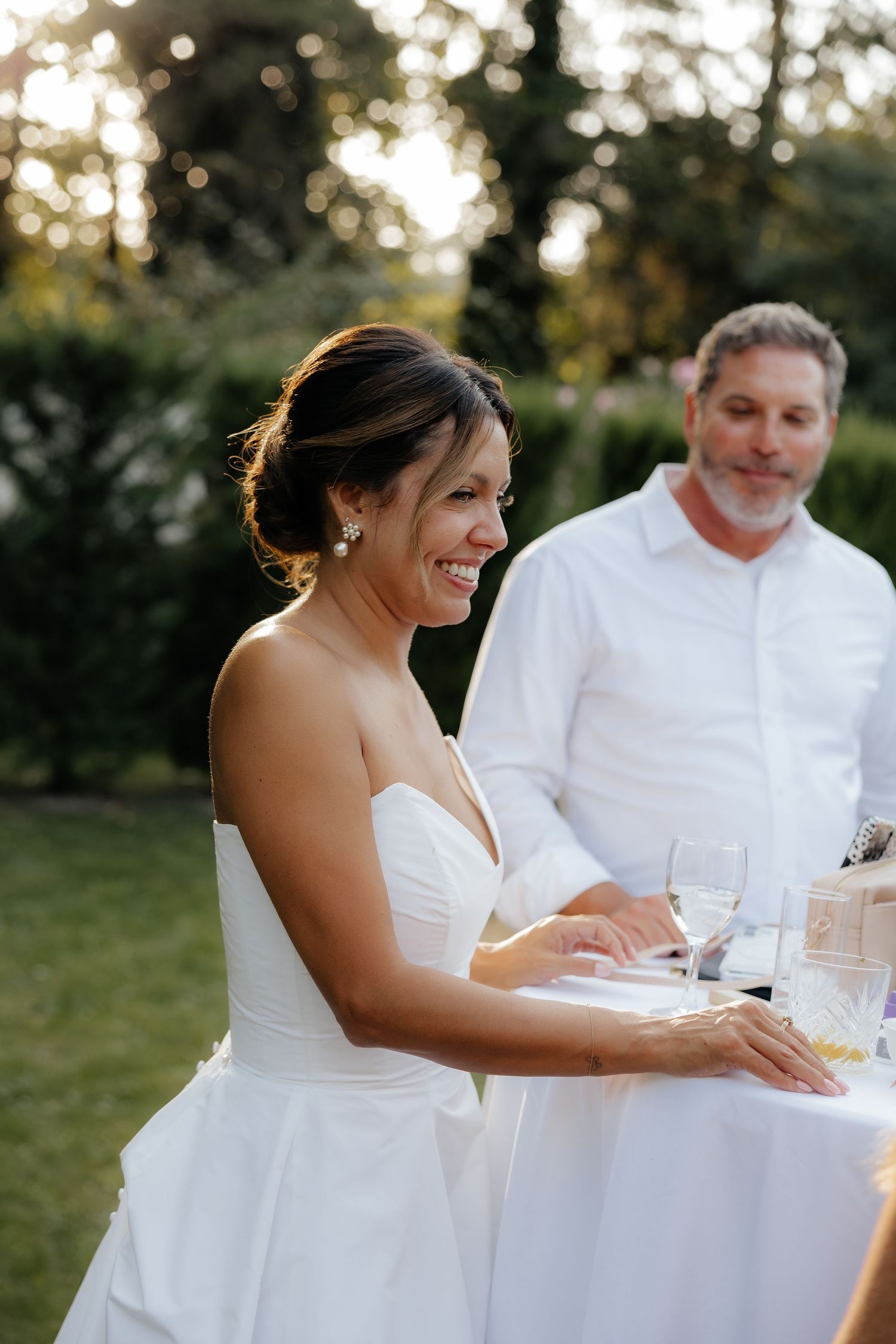 Bride in white dress seated at reception table during golden hour outdoors.