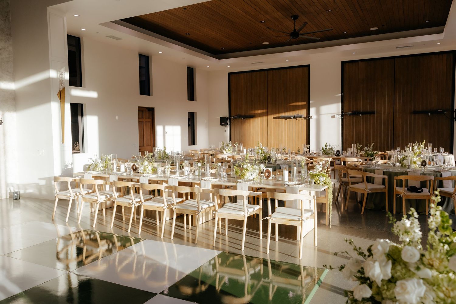 Sunlit wedding reception venue with long wooden tables, light wood chairs, and white floral arrangements.