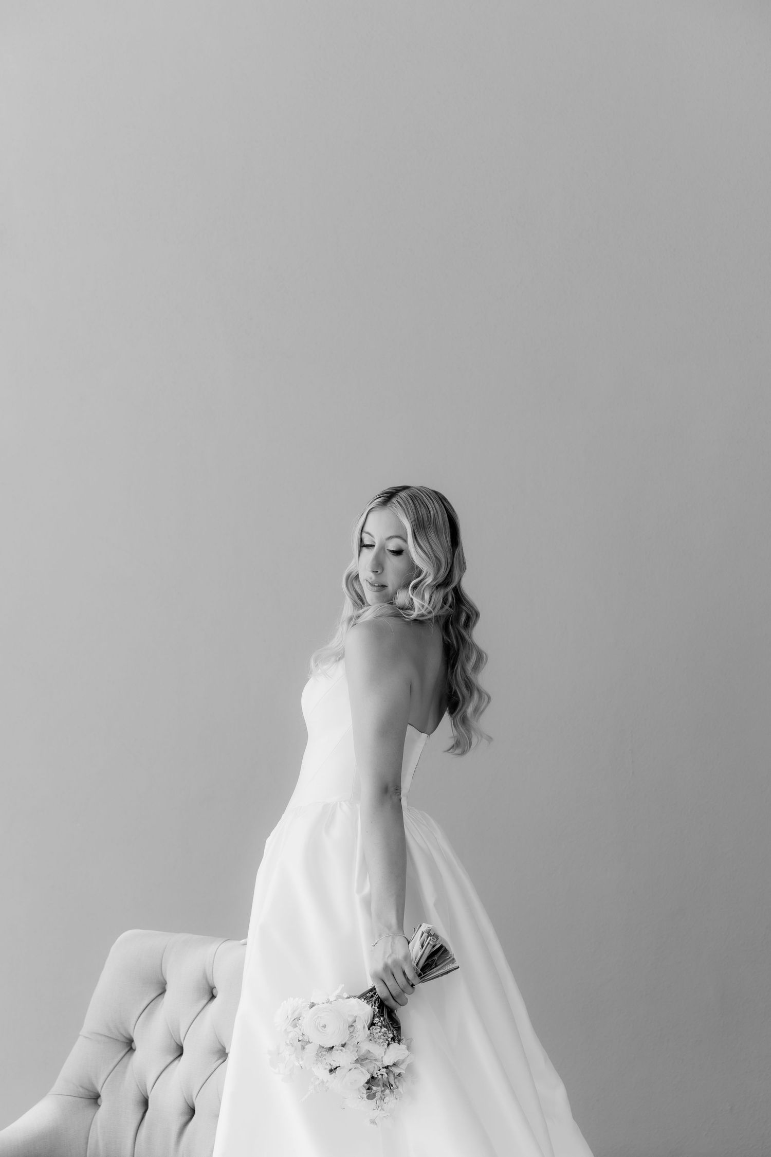 Black and white portrait of a bride in a strapless wedding gown holding a bouquet next to an elegant tufted chair.