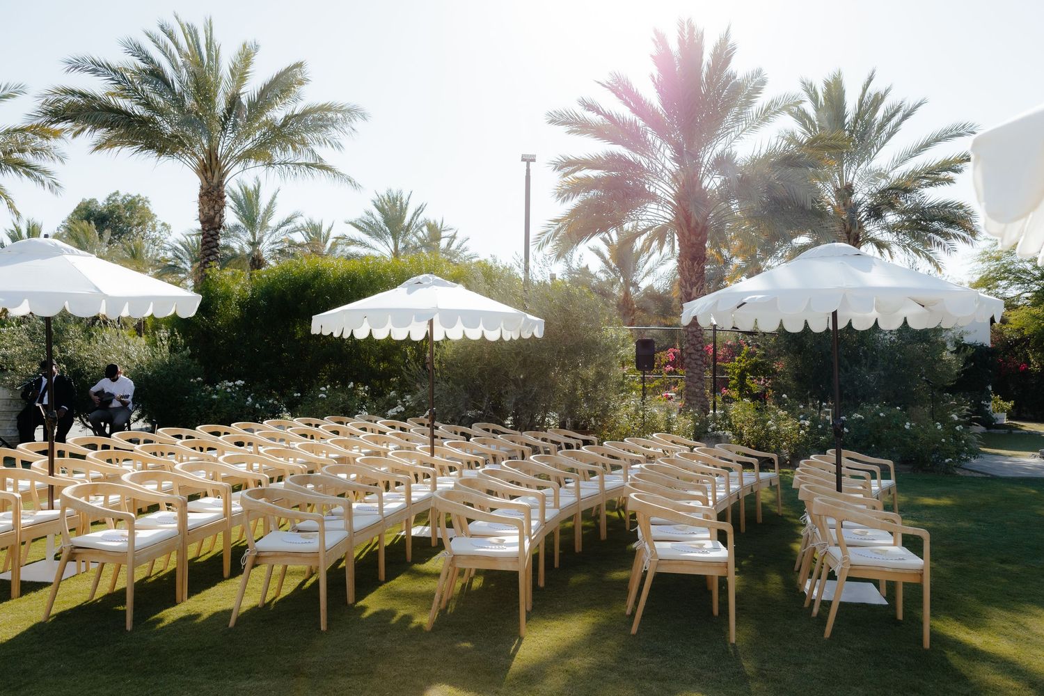 Outdoor wedding ceremony setup with wooden chairs, white umbrellas and palm trees in sunlight.
