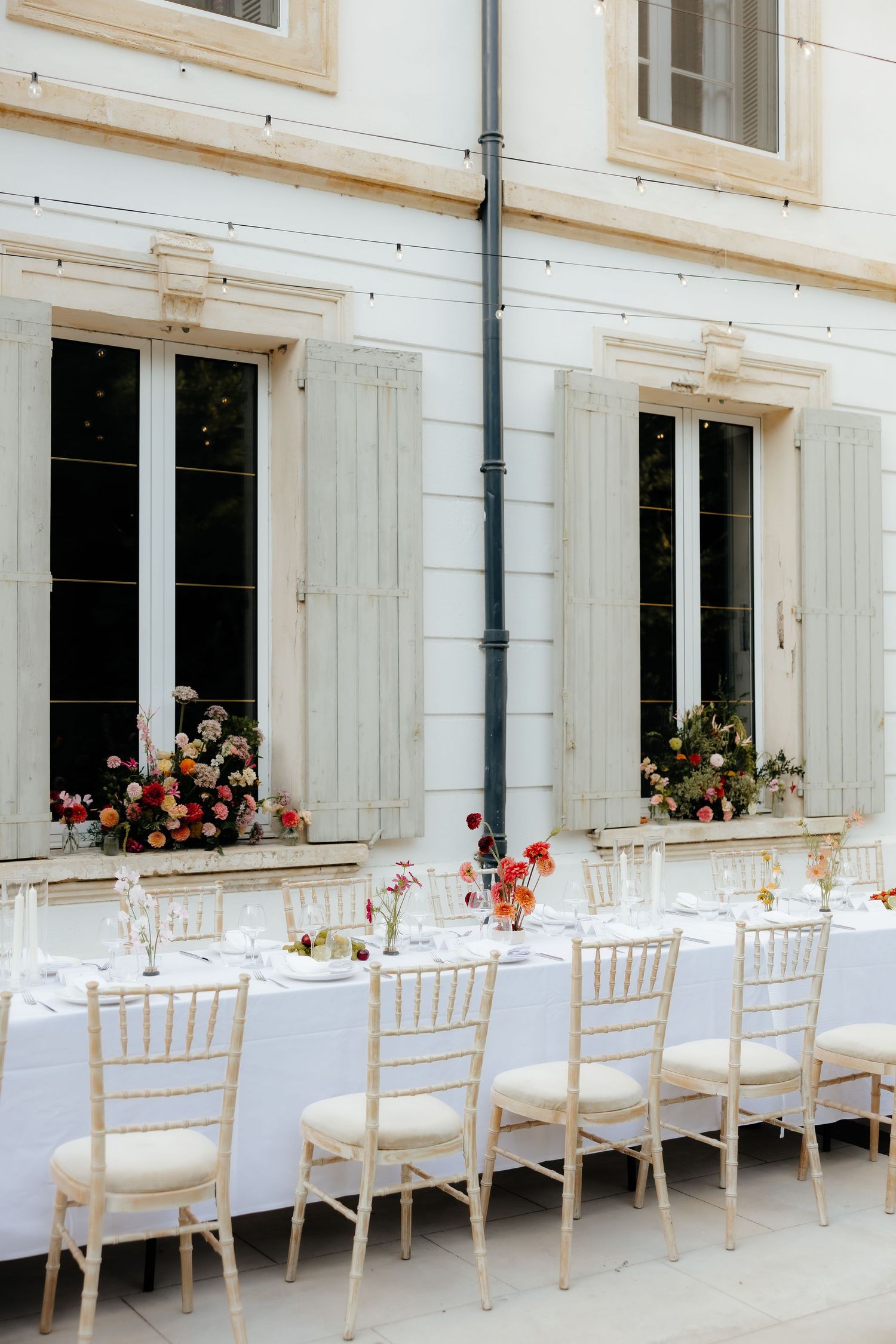 Long elegant wedding reception table setup with white linens and chiavari chairs against a white building exterior.