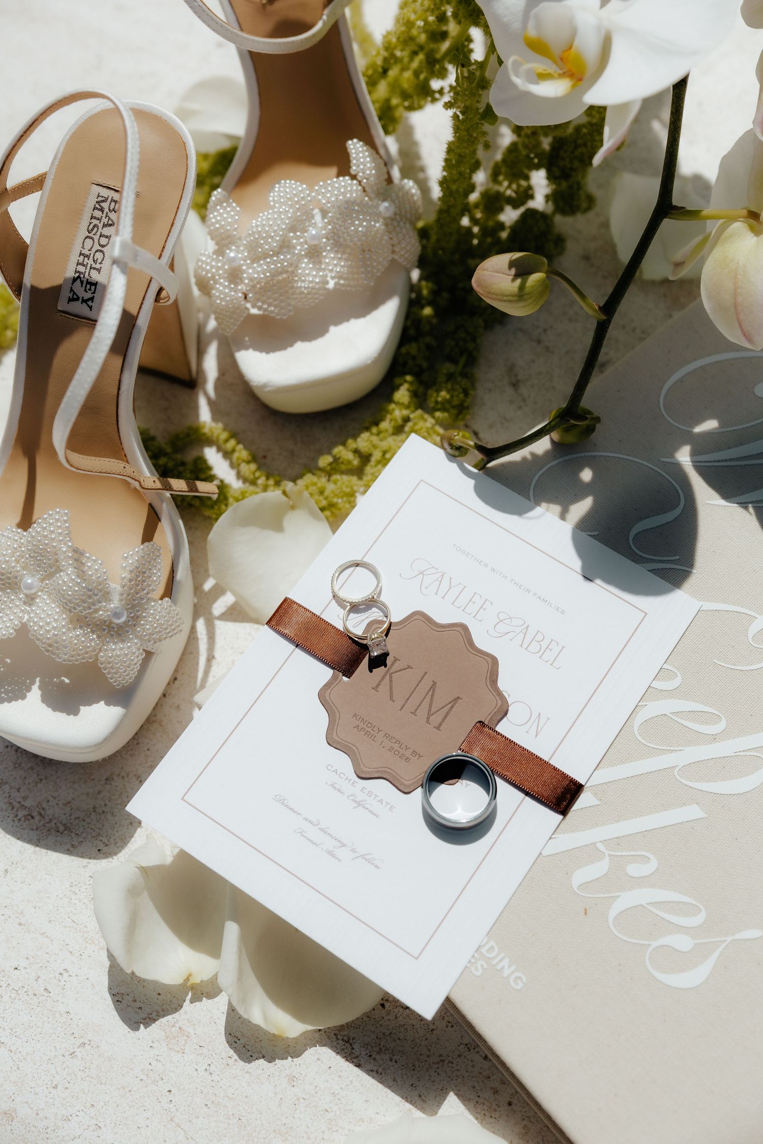 White pearl wedding heels, floral bouquet, gold rings, and elegant wedding invitation styled in a bridal flat lay.