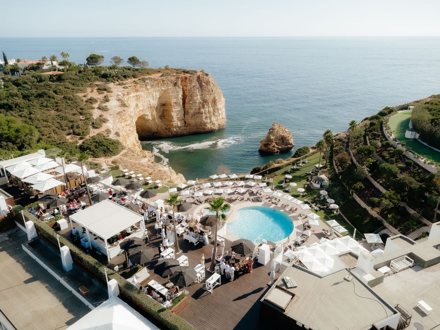 Aerial view of a luxury resort pool with white umbrellas overlooking dramatic cliffs and turquoise sea in the Algarve, Portugal.