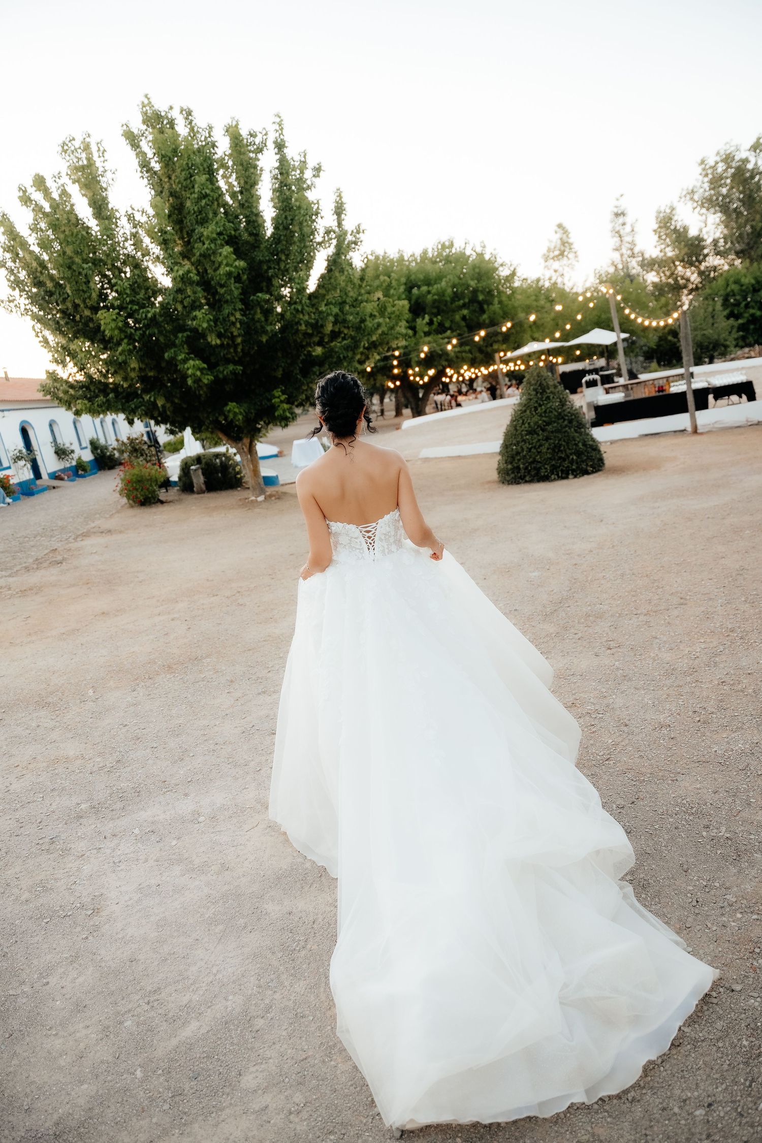 Bride in strapless layered white ballgown walking through rustic outdoor venue with string lights at dusk.