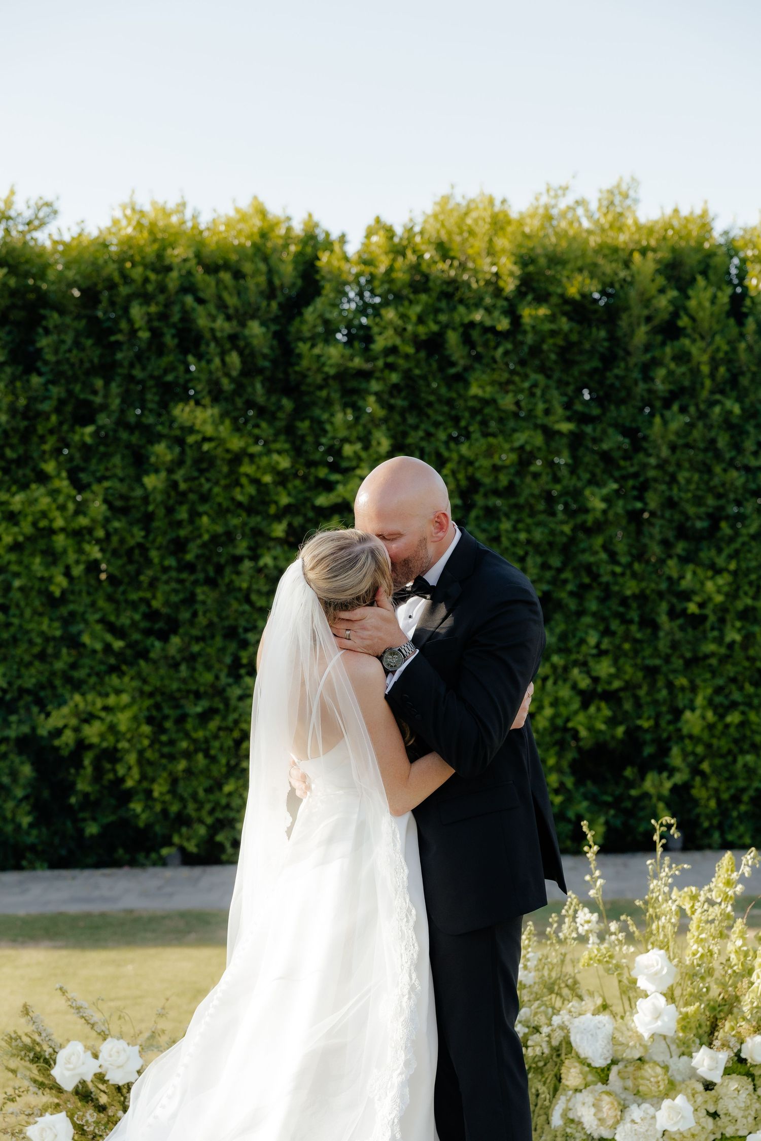 Bride and groom share their first kiss during an outdoor wedding ceremony with lush green hedge backdrop.