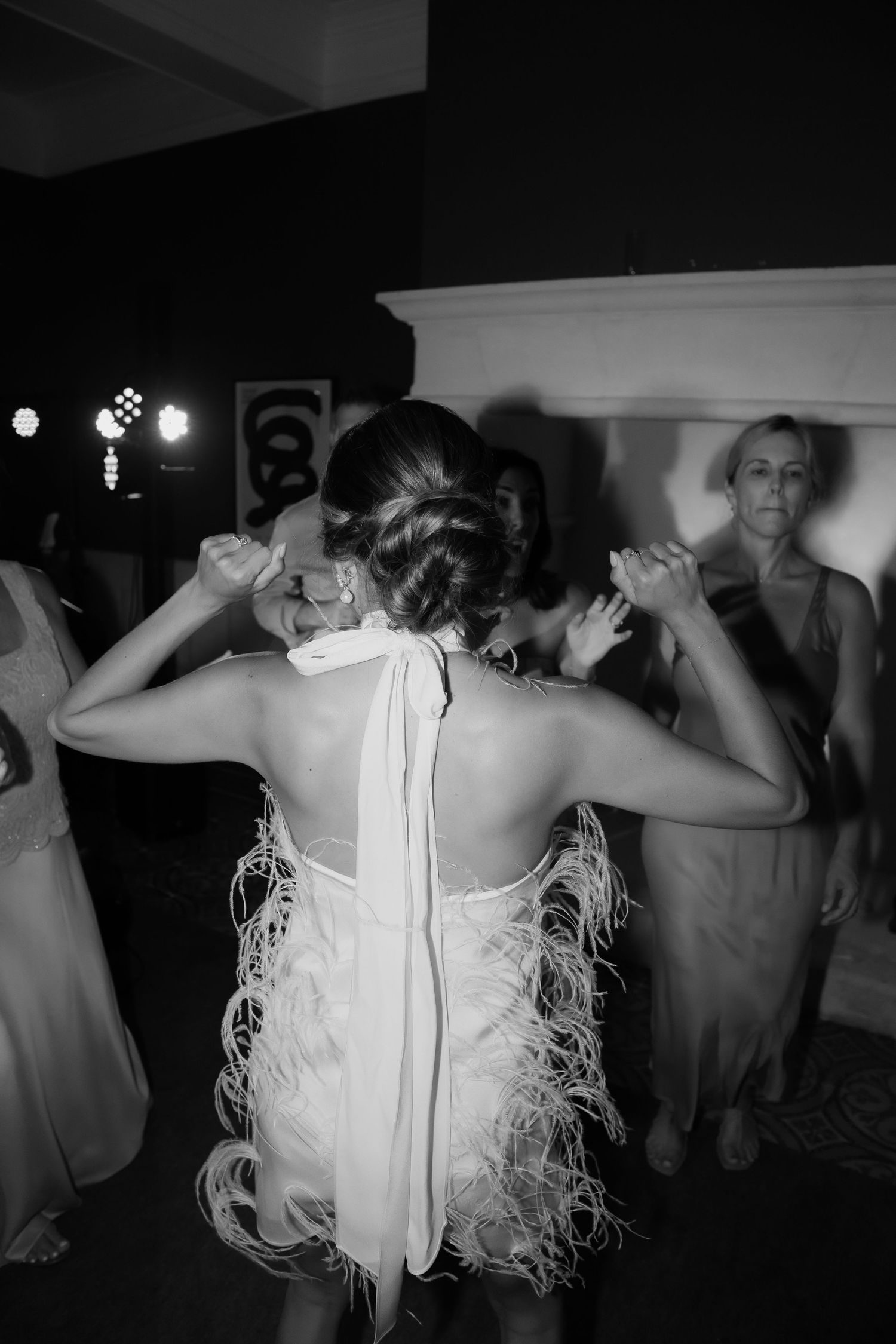 A person in a white fringed dress dances with arms raised on a dark dance floor at a party.