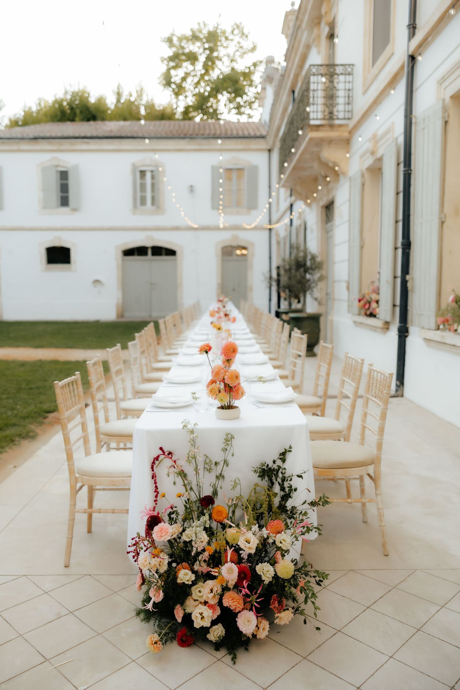 Elegant outdoor wedding reception setup with long tables, white chairs, and colorful floral centerpieces under a covered walkway.