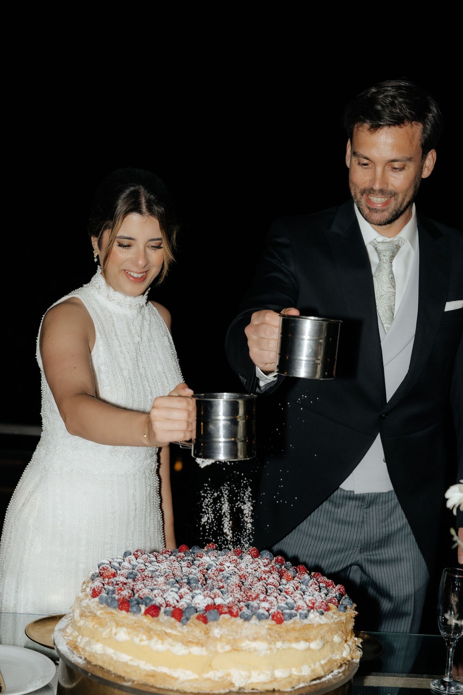 Bride and groom dusting powdered sugar over berry-topped wedding cake at reception.