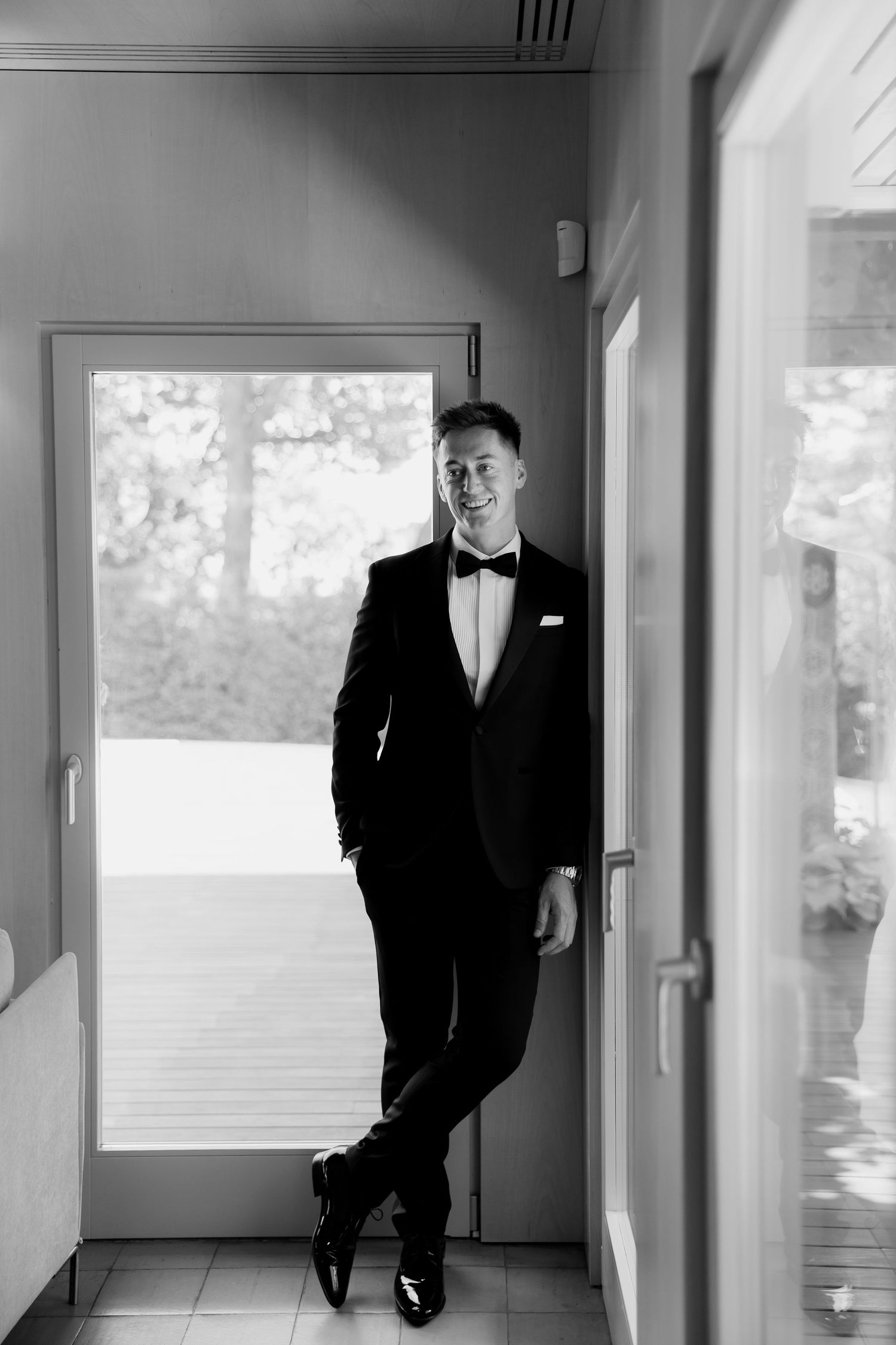 Smiling groom in black tuxedo with bow tie posing in a doorway in a stylish black and white portrait photo.