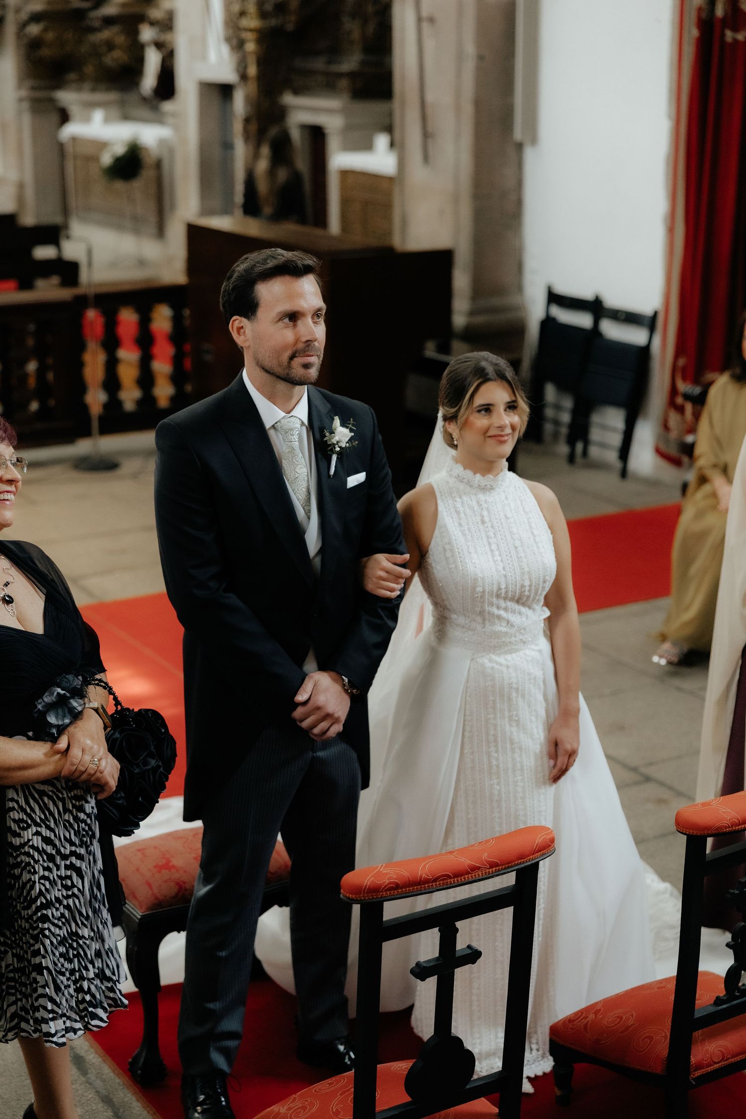 Bride in high-neck white gown and groom in dark suit stand at church altar.