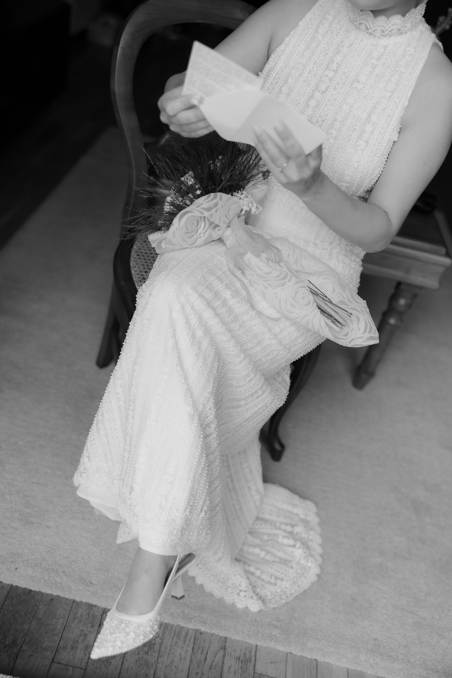 Black and white photo of seated bride in lace gown holding vows with decorative fan bouquet.