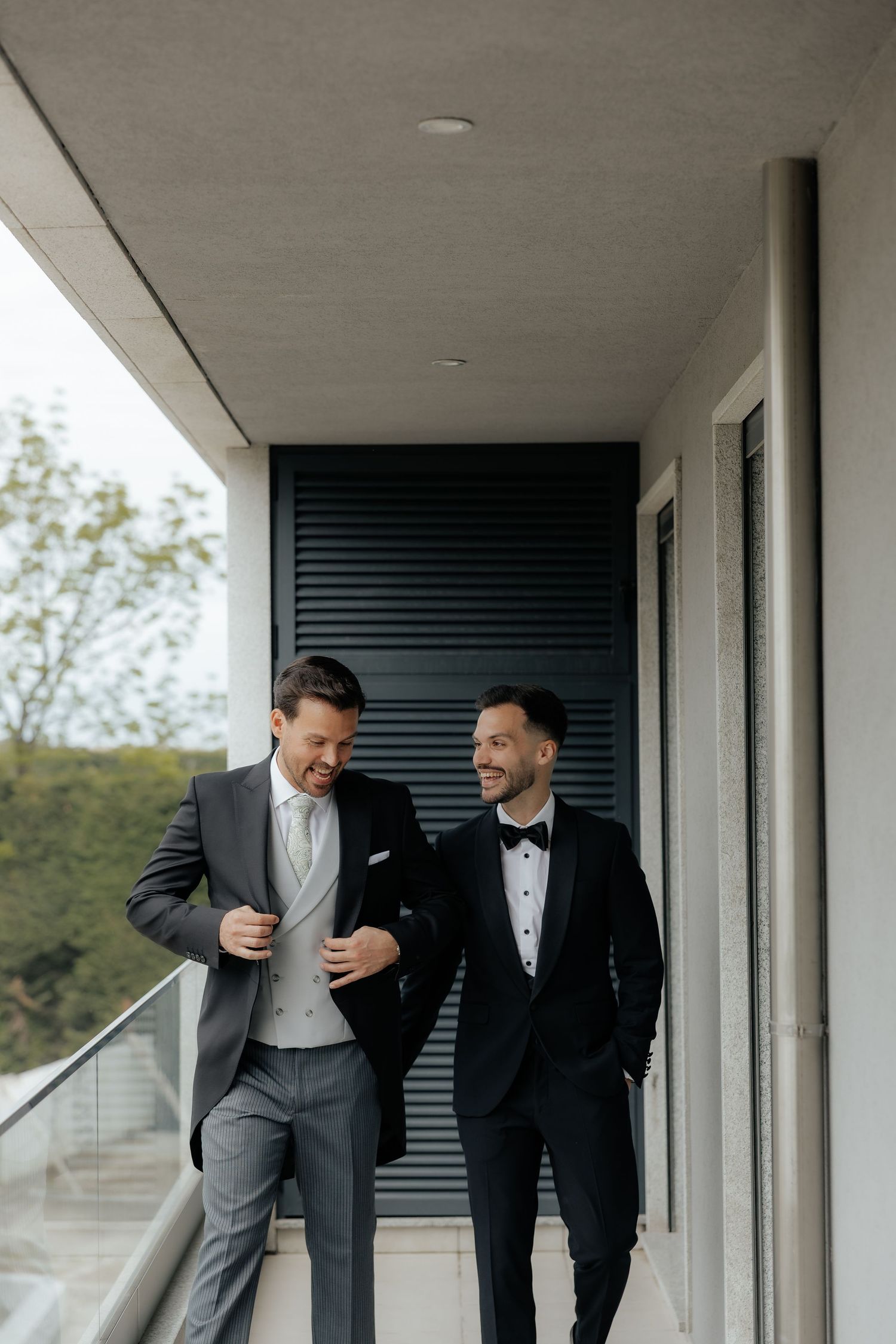 Two men in elegant wedding suits smile and walk together on a contemporary outdoor balcony.