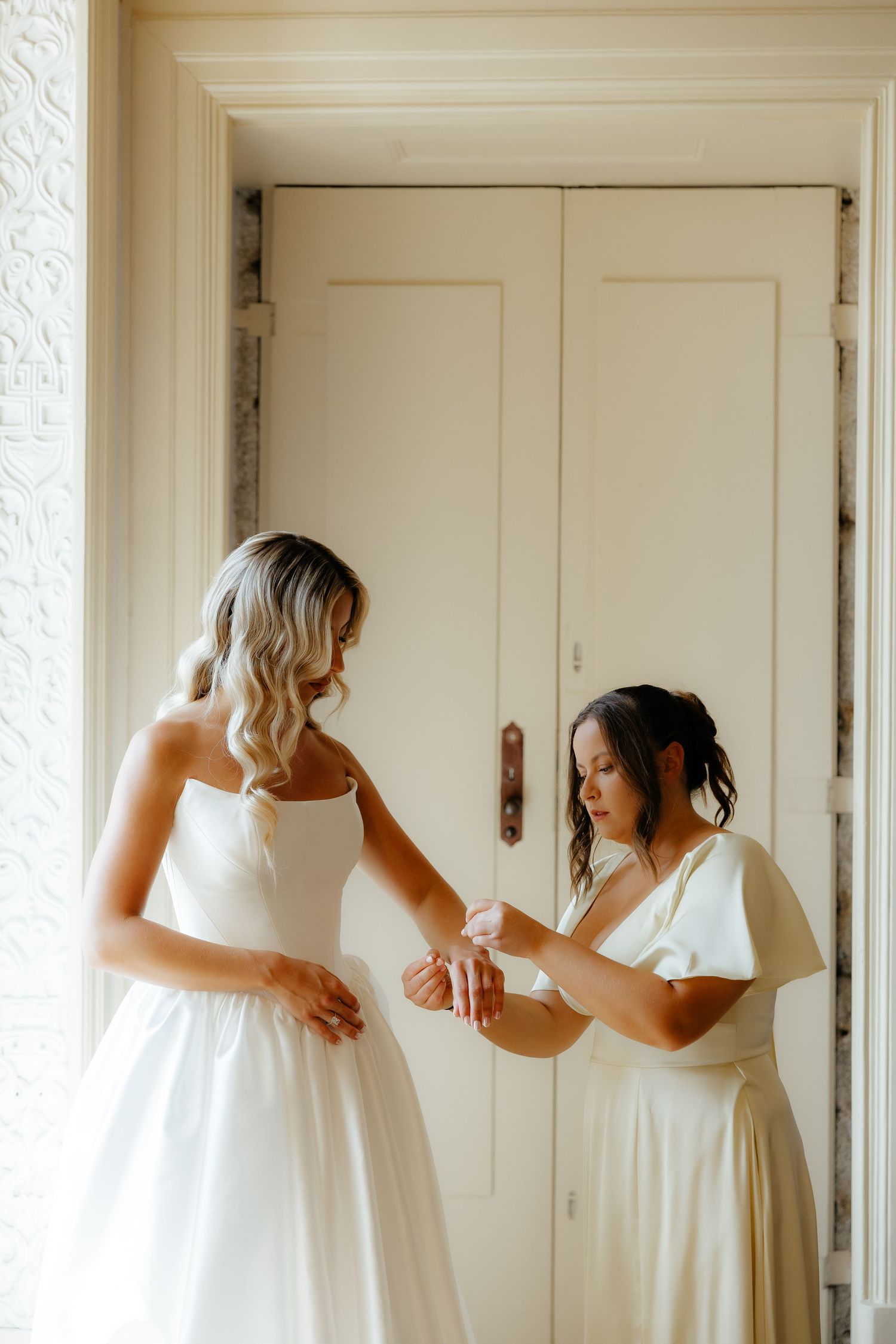 Bridesmaid in white dress helping bride with bracelet in front of classic double doors.