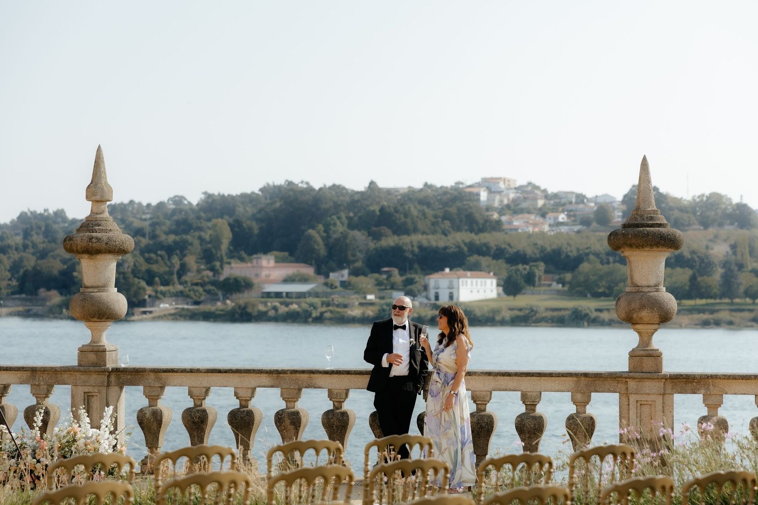 Elegant couple standing on ornate stone balcony overlooking scenic waterfront at a luxury wedding venue.