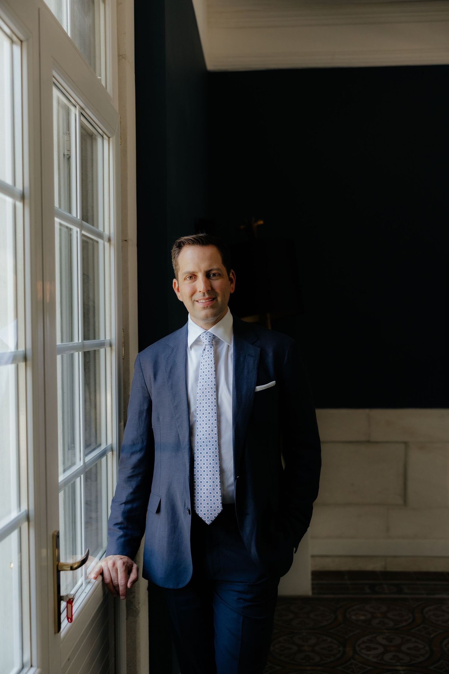 Elegant groom in blue suit and tie standing by large windows in natural light.