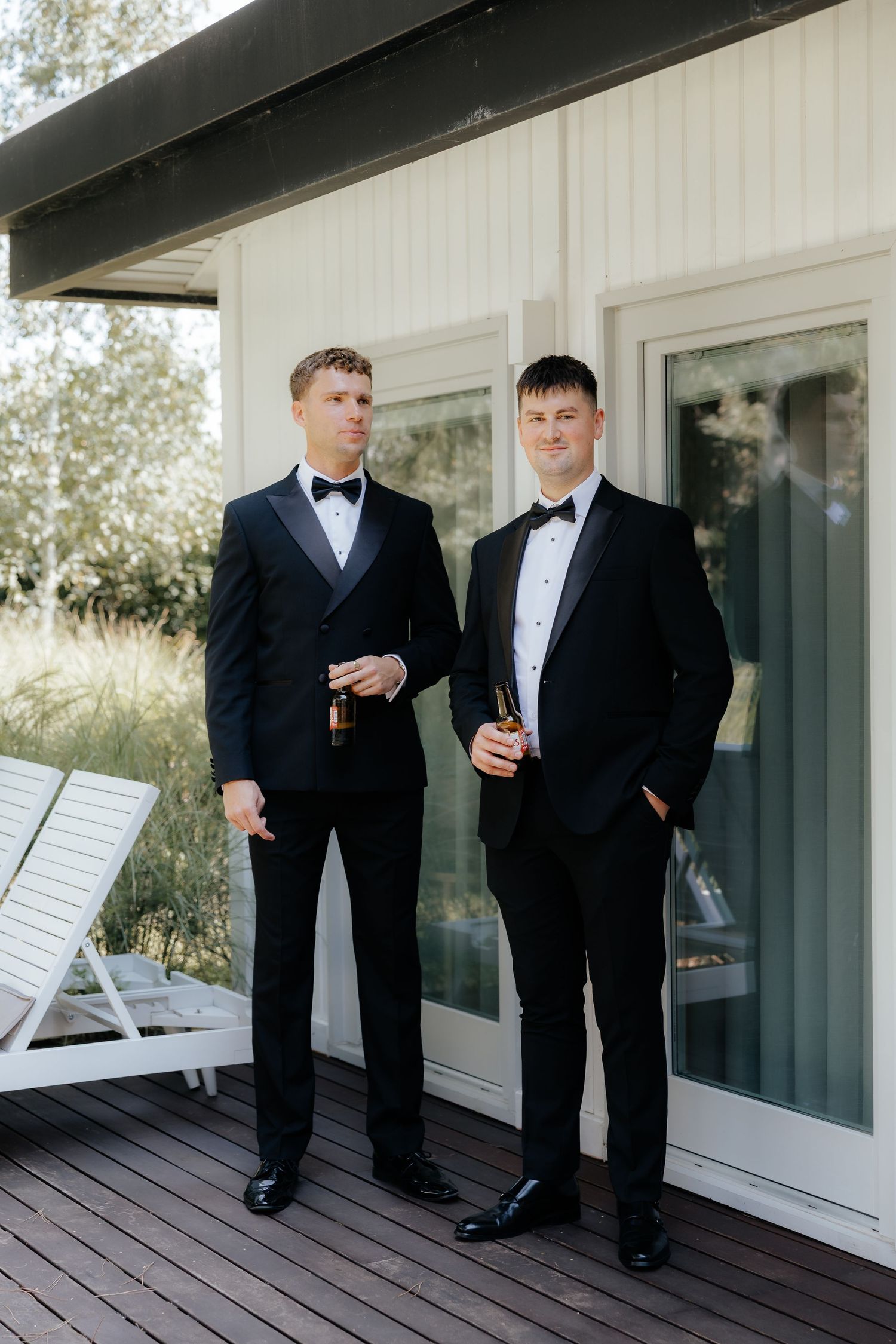 Two men in black tuxedos and bowties stand together on a white deck outside a building.