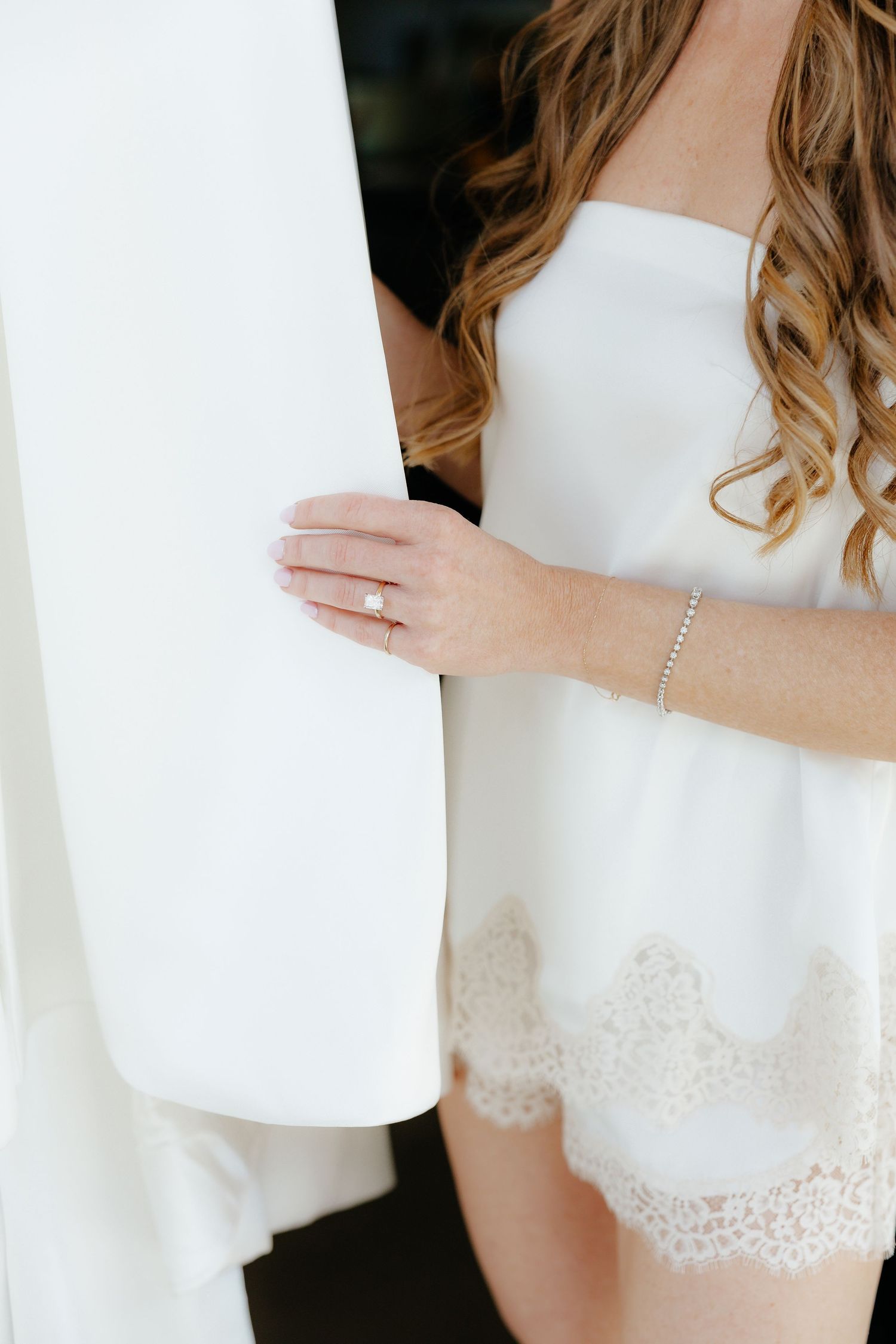 Close-up of bride in white lace shorts holding wedding dress with diamond engagement ring visible.