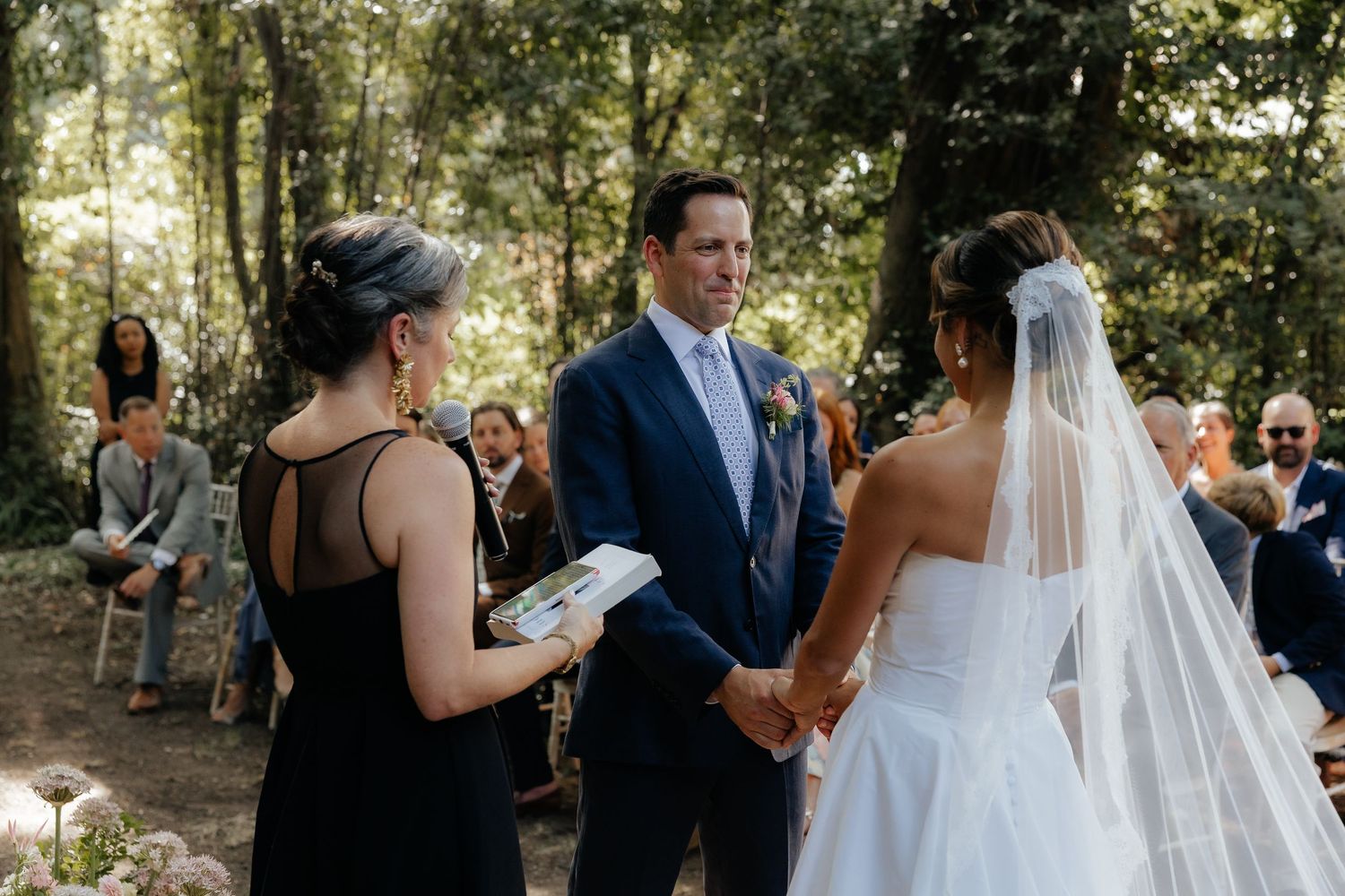 Outdoor wedding ceremony in a wooded setting with guests watching the couple exchange vows.