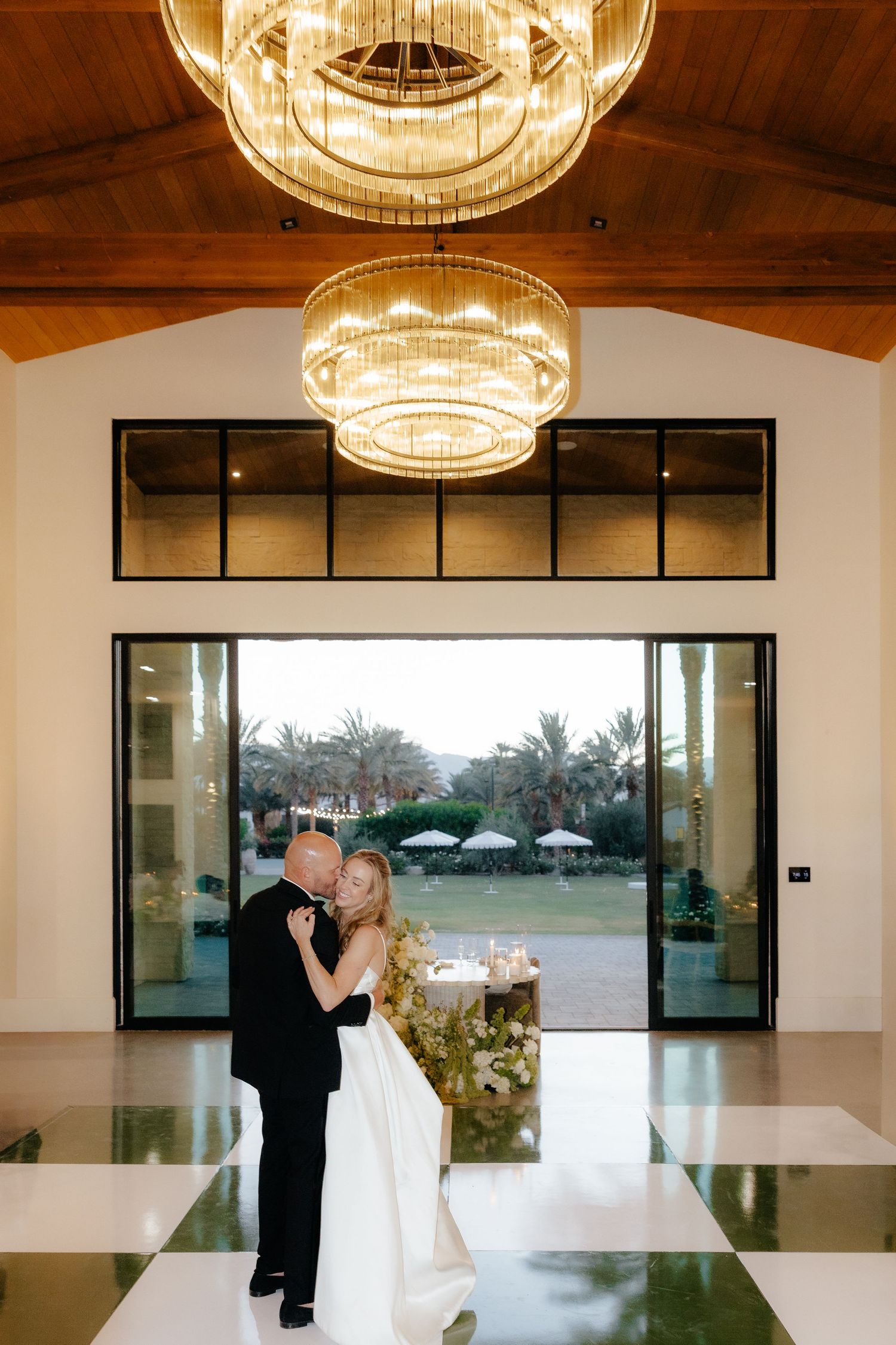 Bride and groom share a romantic kiss on a checkered dance floor beneath elegant gold chandeliers at their wedding reception.