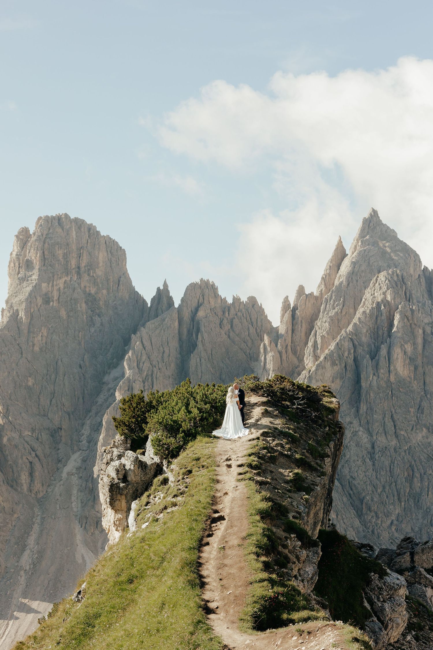 Bride in white gown standing on rocky mountain ridge in the Dolomites with dramatic peaks behind.