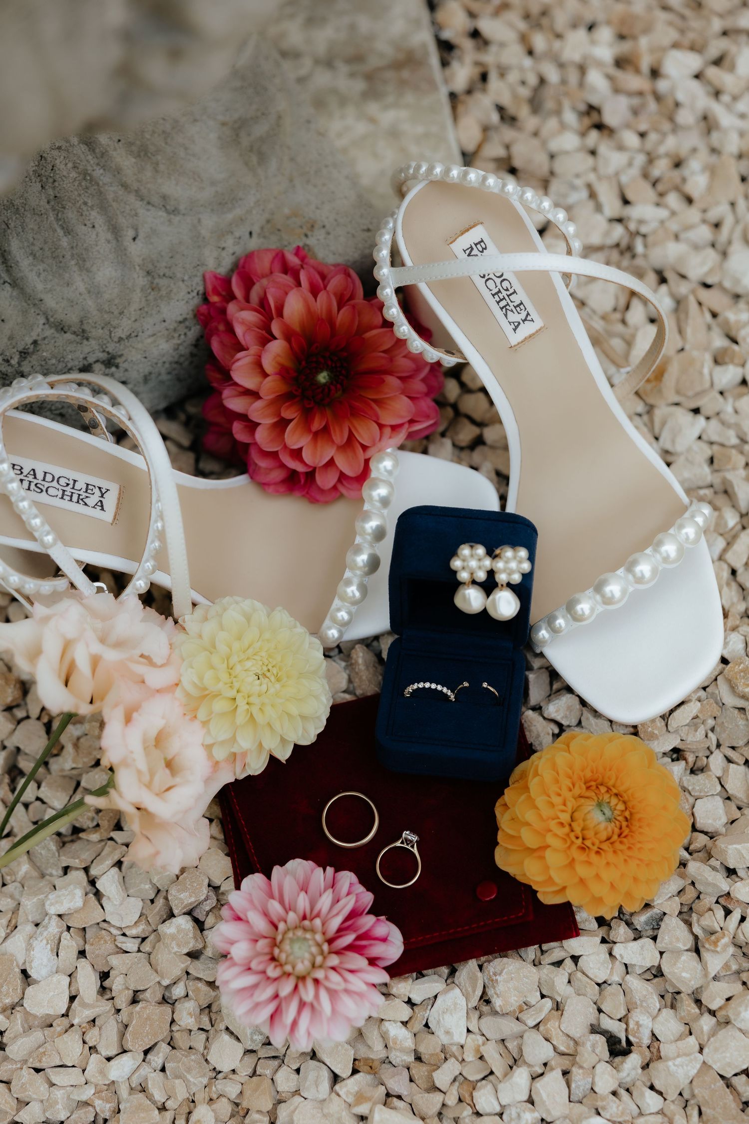Wedding accessories including white pearl sandals, colorful dahlias, and jewelry arranged on a textured surface.