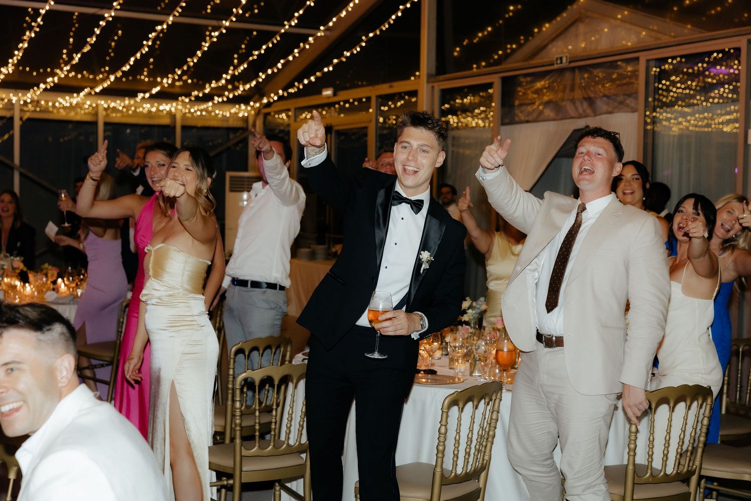 Wedding guests dancing and celebrating at a reception under string lights in an indoor venue.