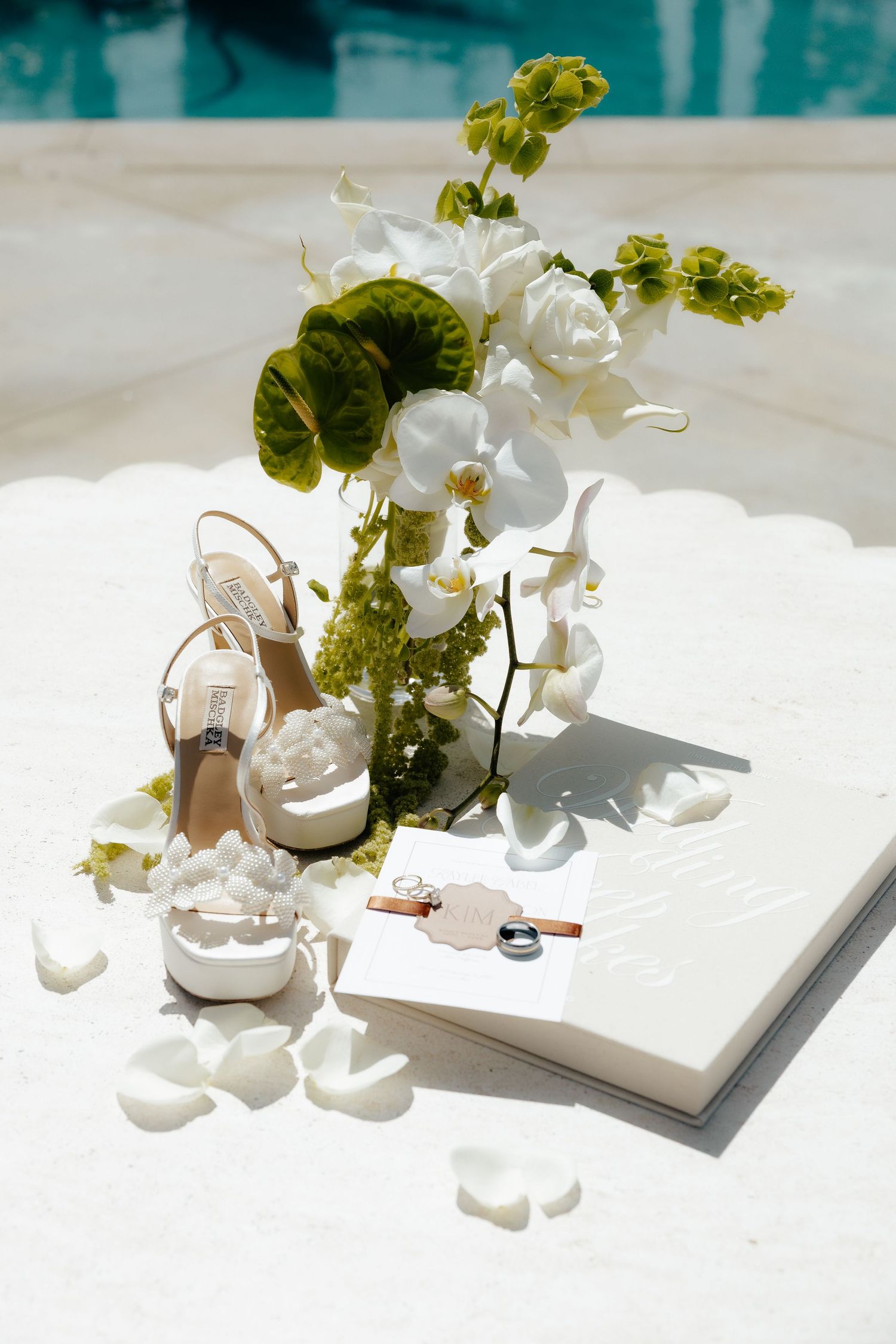 Wedding flat lay with pearl heels, white orchids, vow book, and rings by poolside.