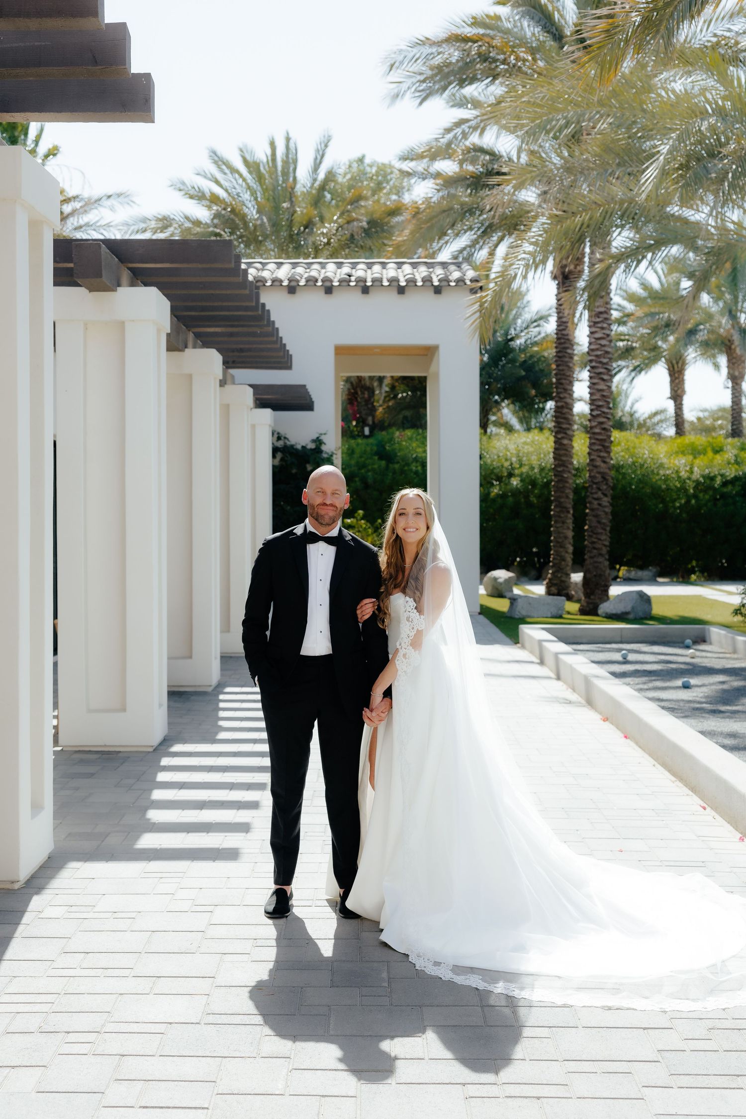 Bride and groom walking together along a sunlit outdoor colonnade lined with palm trees.