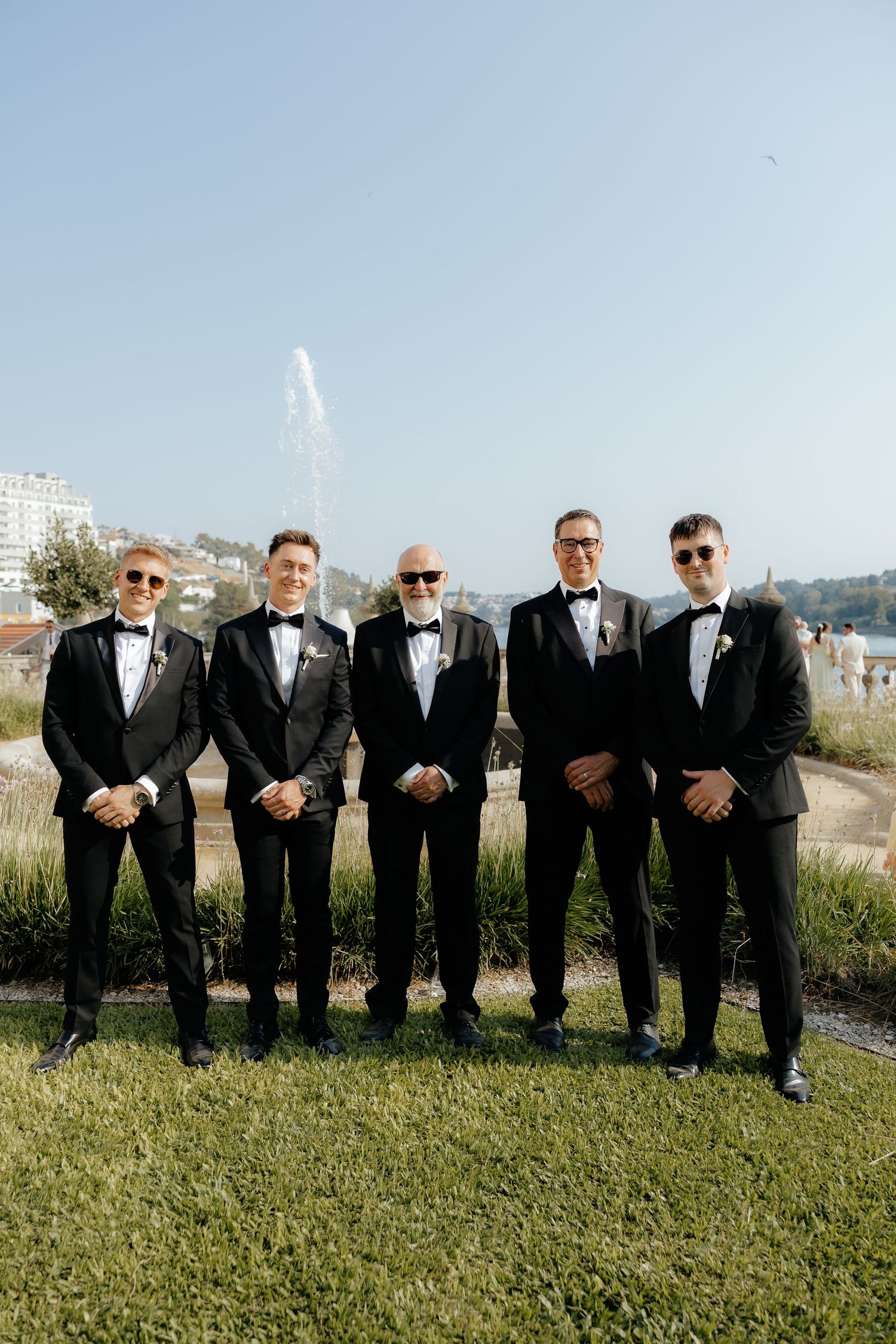 A row of groomsmen in black tuxedos and sunglasses pose outdoors on grass against a scenic backdrop.
