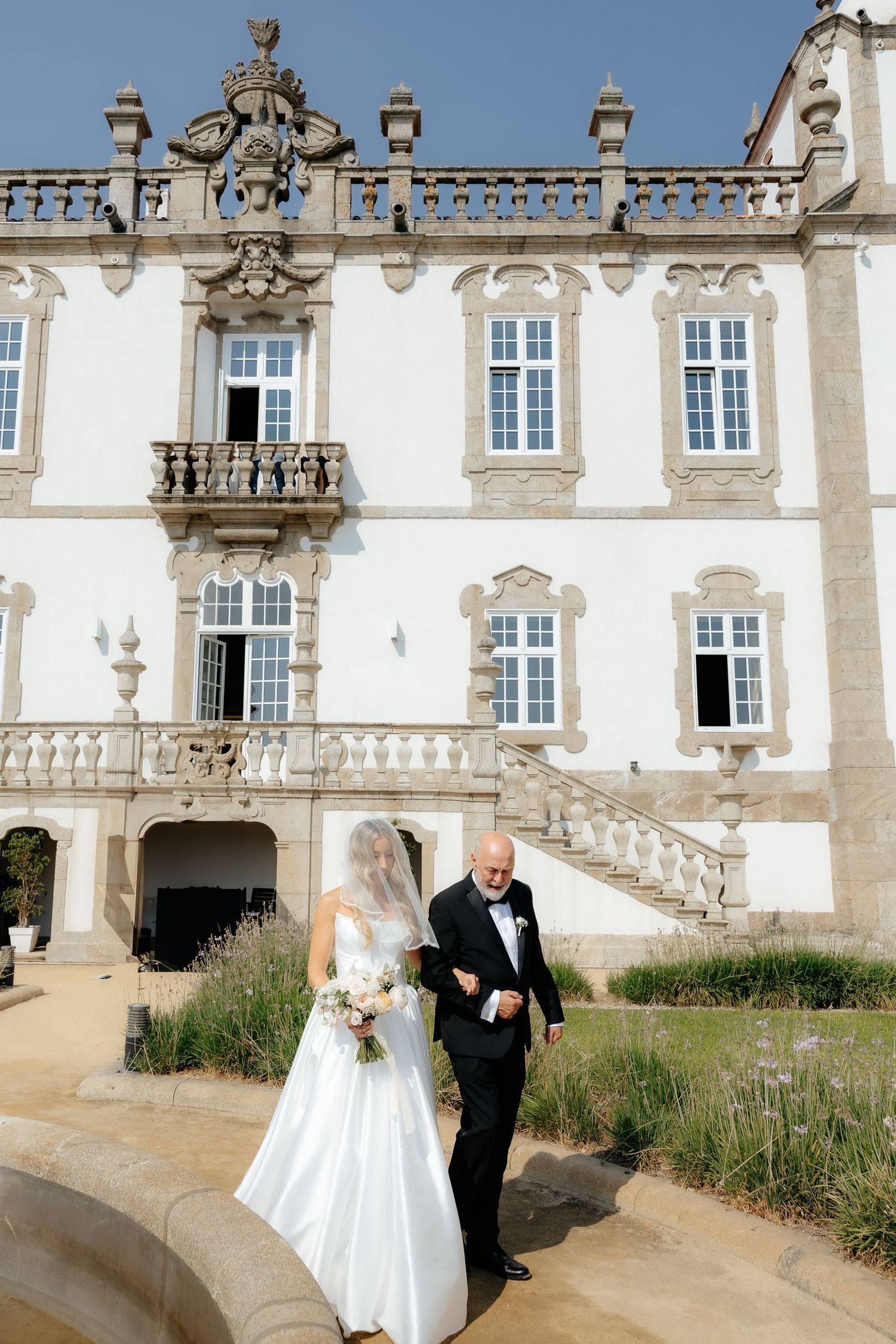 Elegant couple in wedding attire walking down steps of ornate historic white mansion with decorative balustrades and windows.