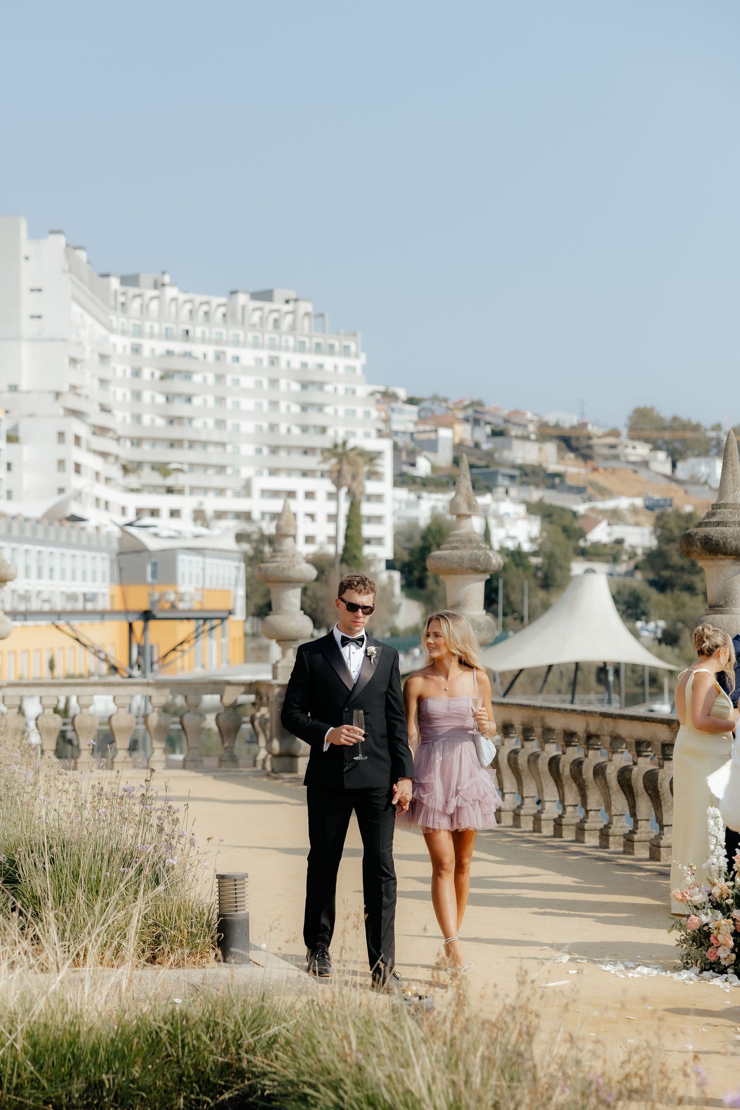 A couple walks along a scenic balustrade overlooking a coastal hillside with white buildings and blue sky in Portugal.