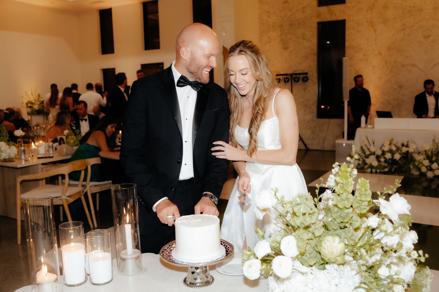 Smiling bride and groom cut their simple white single-tier wedding cake surrounded by white floral arrangements and candles.