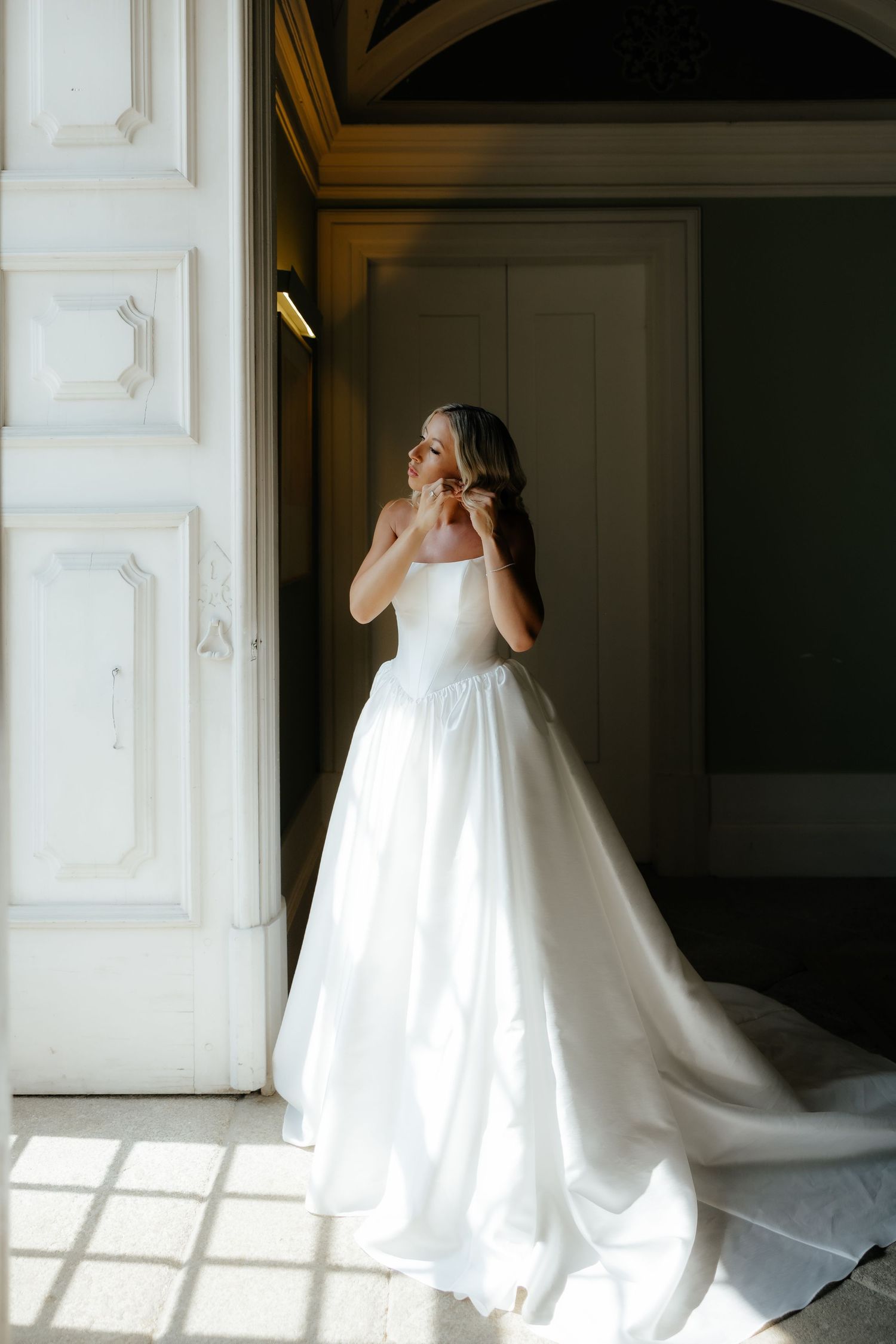 A bride in a flowing white wedding gown poses gracefully in front of large sunlit windows in an elegant interior space.