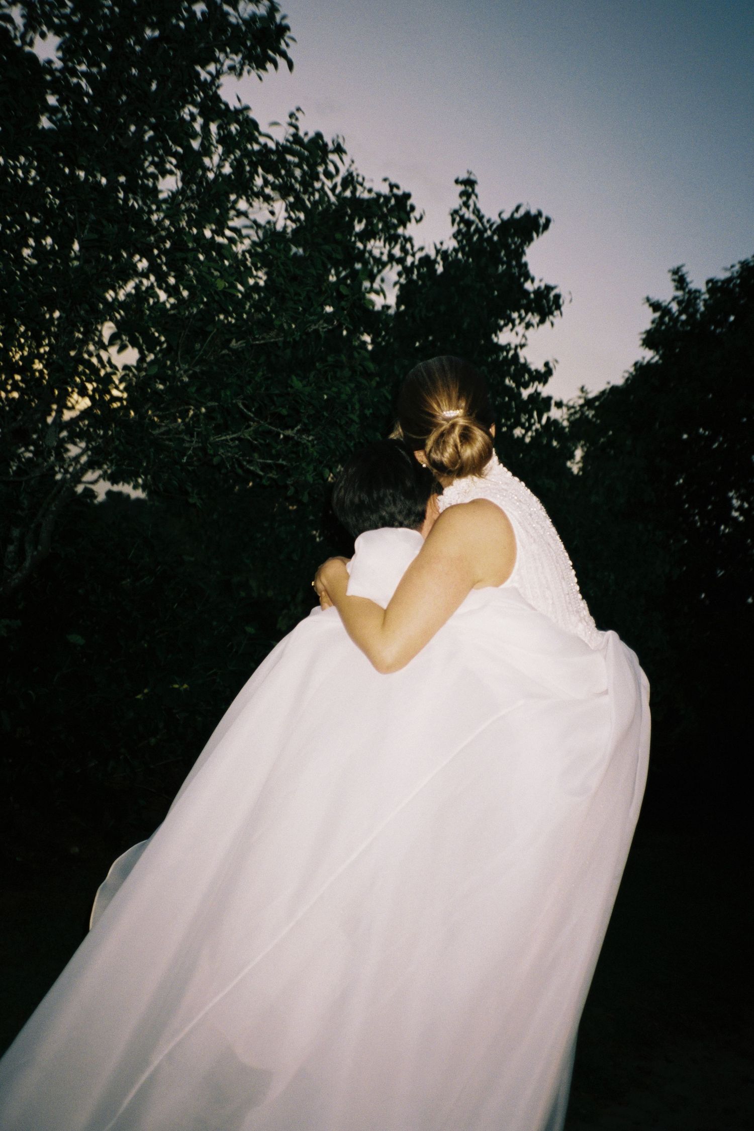 Bride and bridesmaid share an emotional embrace in a white wedding gown outdoors at dusk.