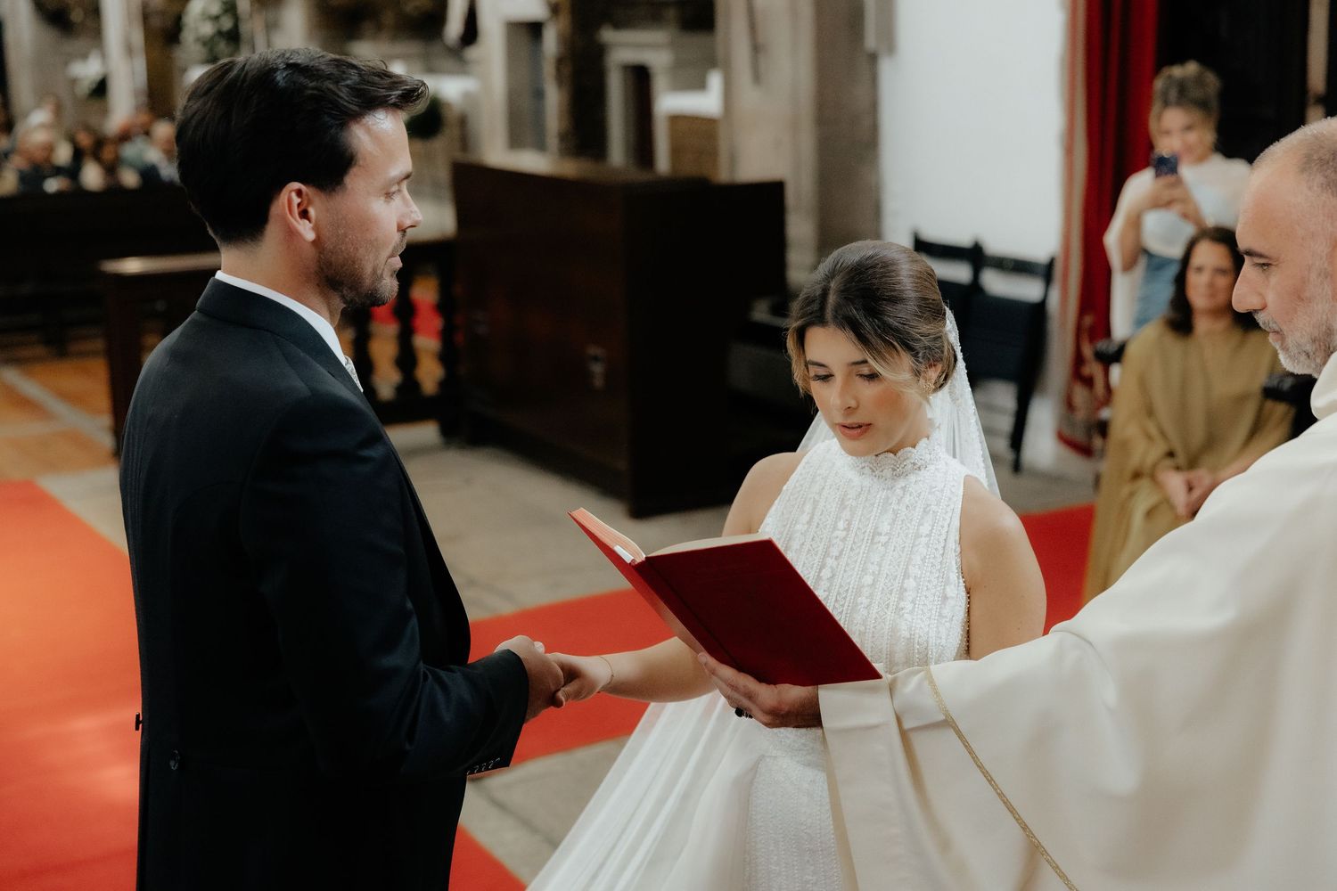 Bride and groom exchange vows holding red book before priest at church wedding.