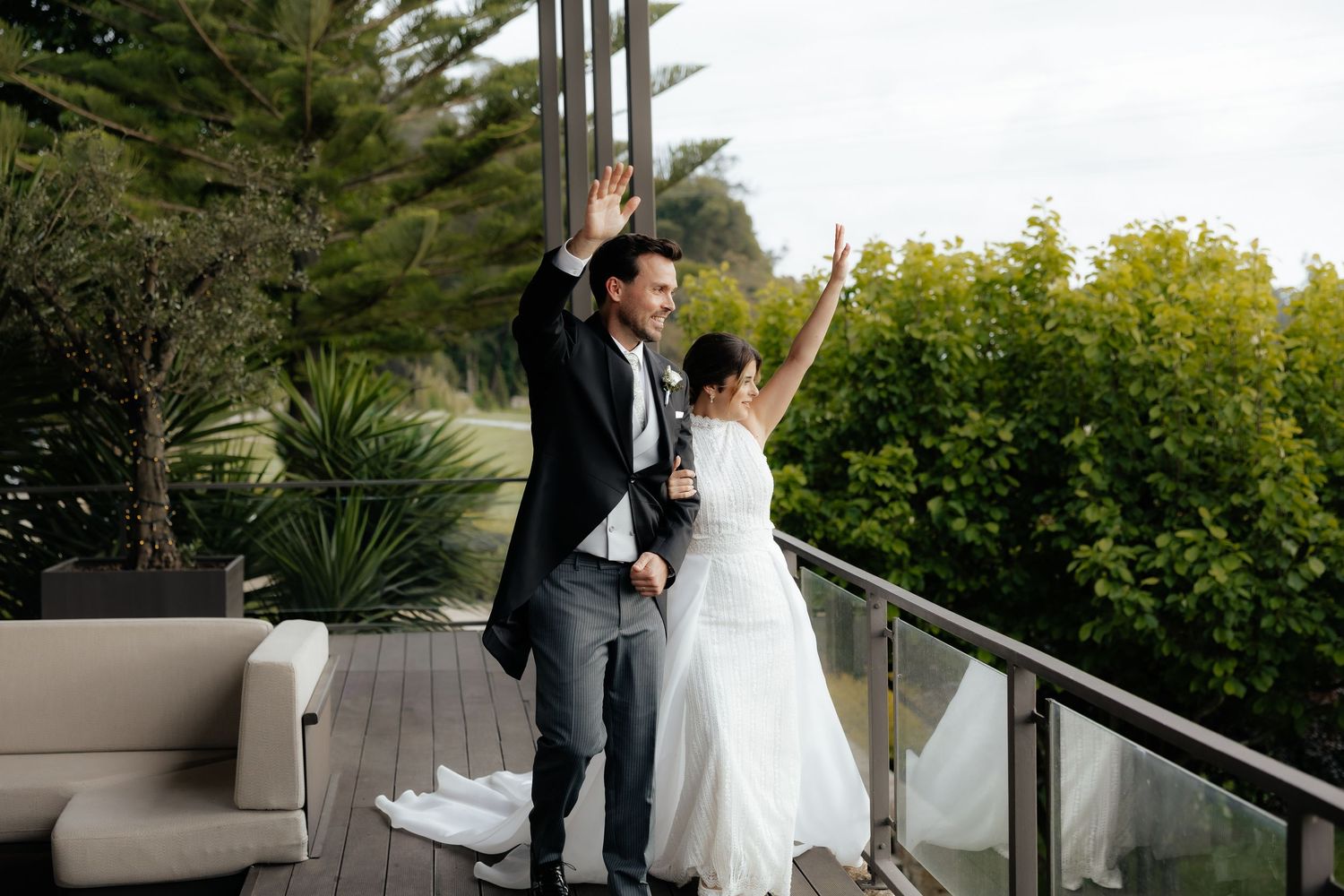 Joyful newlyweds raise arms in celebration on outdoor deck terrace with lush green garden backdrop.