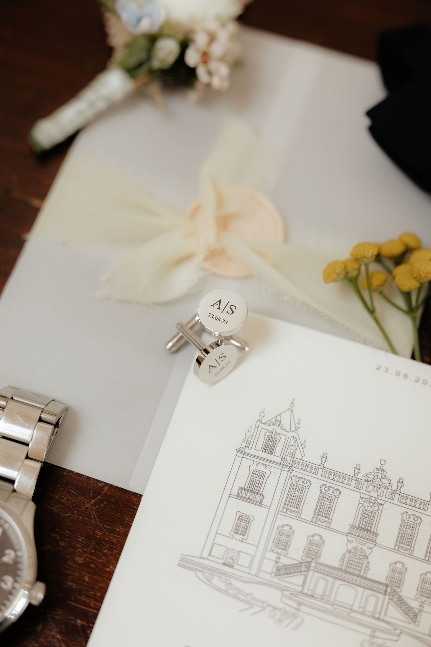 Close-up of monogrammed AJS silver cufflinks resting on a wedding invitation with an illustrated venue drawing.