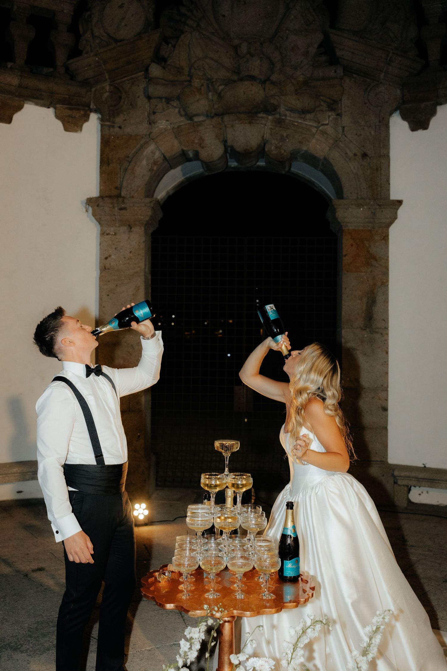 Bride and groom playfully drinking champagne directly from bottles beside their wedding champagne tower.