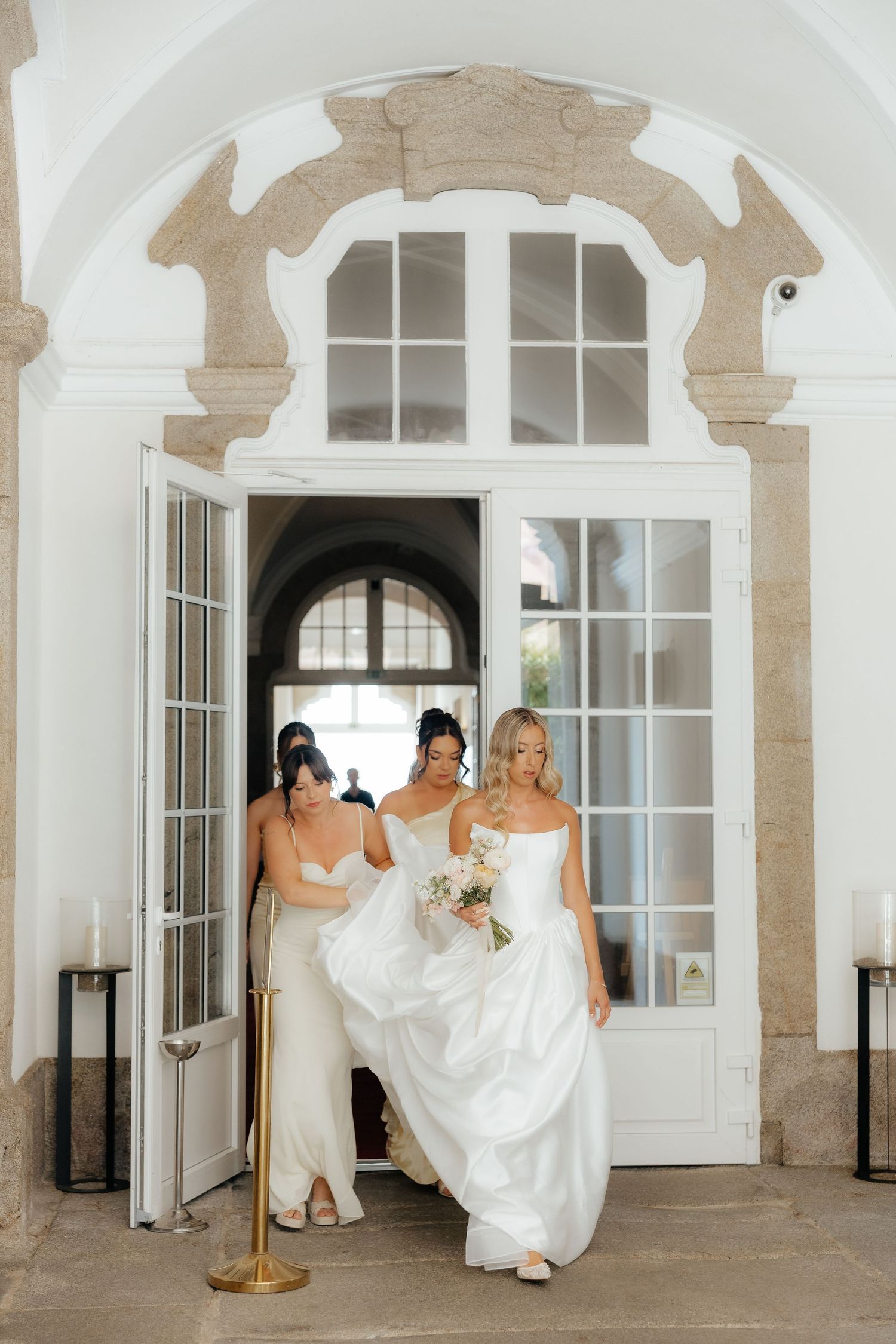 Two bridesmaids help bride in white ball gown exit through ornate arched doorway with candles.